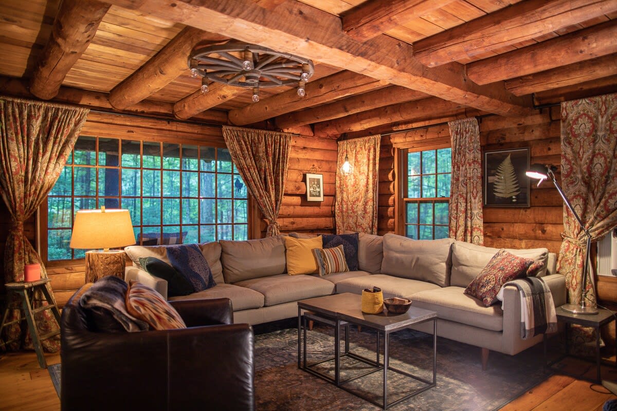 Spacious log cabin living room with a large L-shaped sectional sofa adorned with colorful pillows, rustic wood-beamed ceilings, and multiple windows offering views of the surrounding forest. A wagon wheel chandelier hangs from the ceiling.