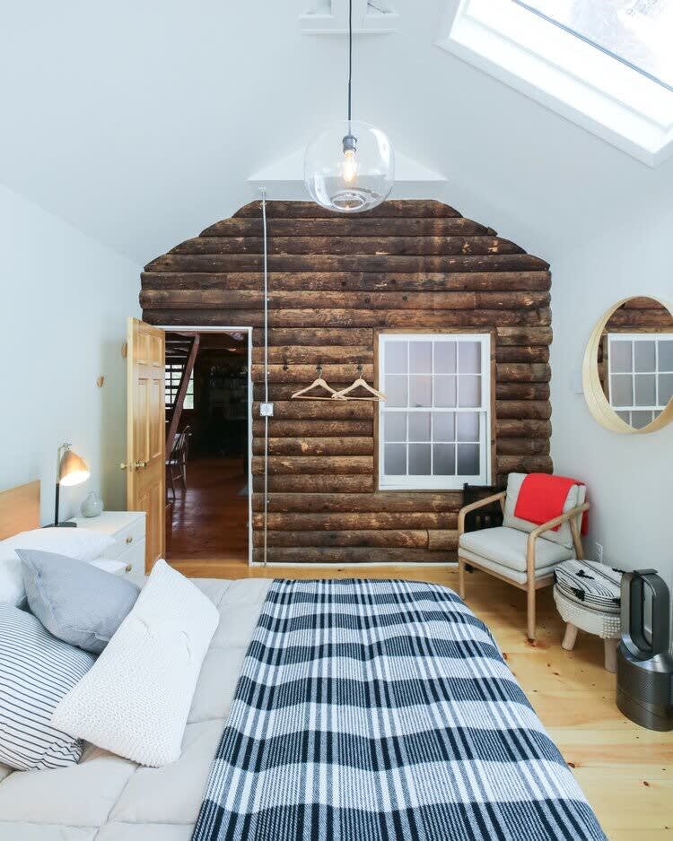 Bright bedroom with a white vaulted ceiling and skylights, featuring a bed with a black and white plaid blanket. One wall is exposed log, creating a cabin feel, while the others are painted white, with a window and a round mirror.