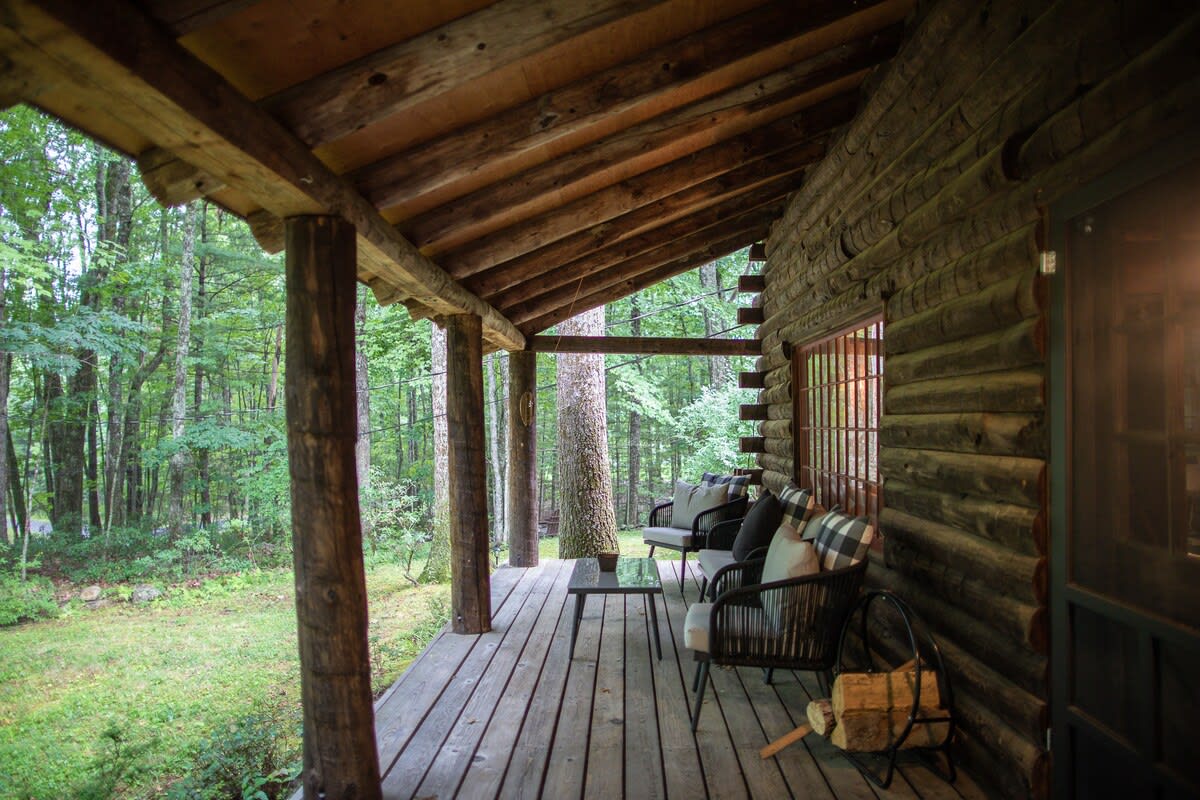 Covered wooden porch with comfortable outdoor seating, including chairs and a bench, overlooking a lush green forest. The porch features rustic log support beams and a wooden floor.