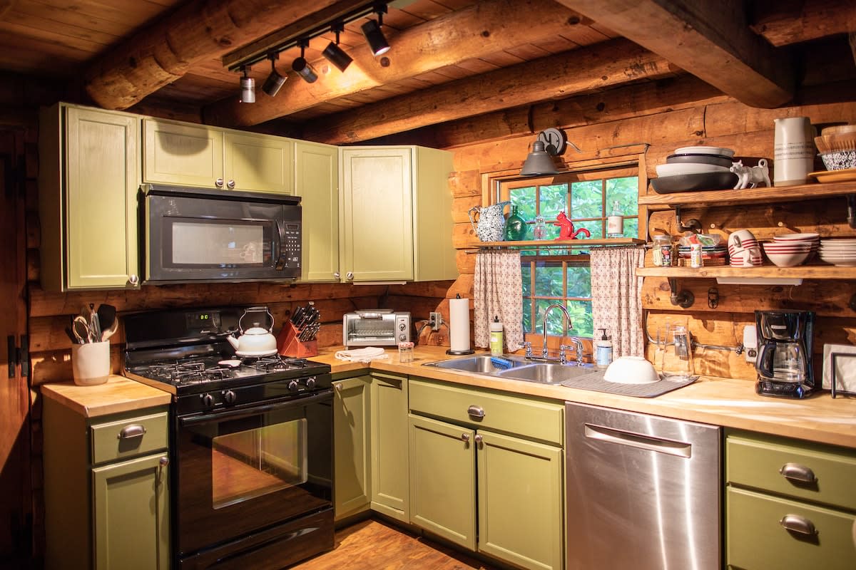 Rustic kitchen with light green cabinetry, black appliances including a microwave and gas range, and light wood countertops. Open shelving displays dishes, and a double sink is positioned under a window with patterned curtains.
