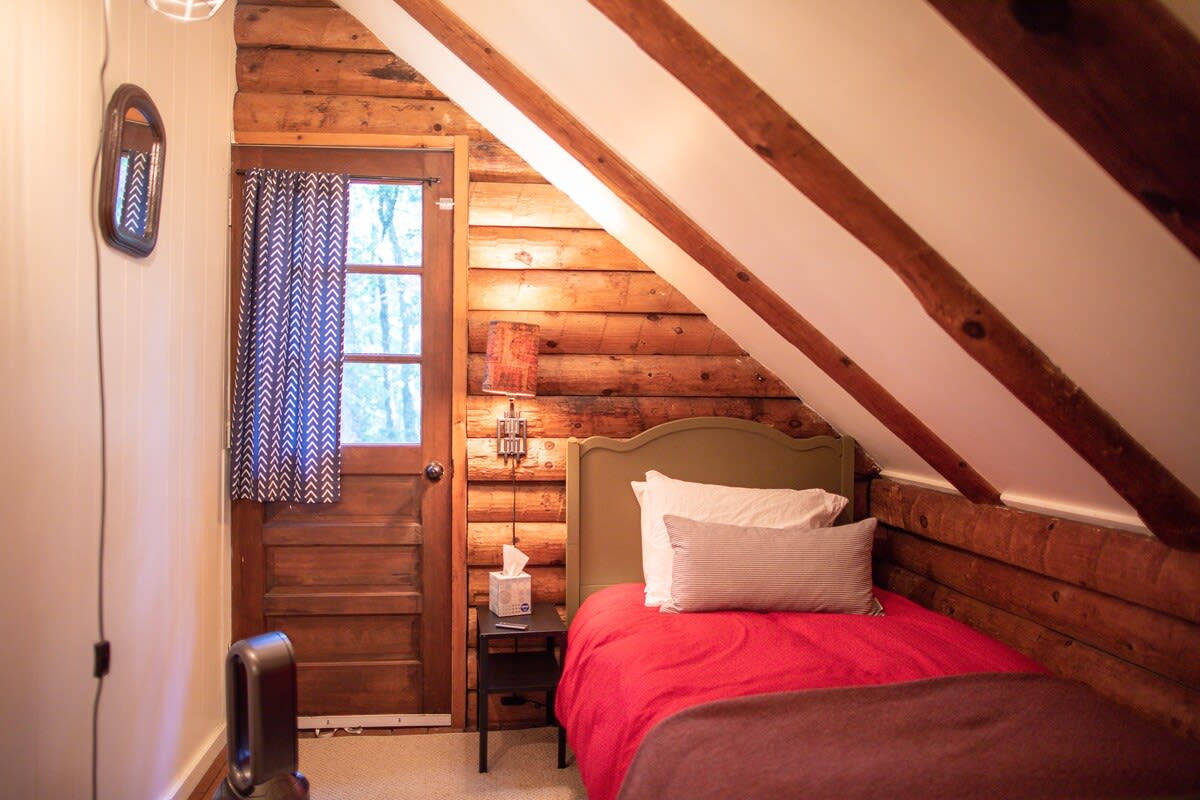 A cozy bedroom with a single bed featuring a red comforter and striped pillow, set against rustic log walls. The room has a sloped white ceiling with exposed wooden beams and a wooden door with a window leading outside.