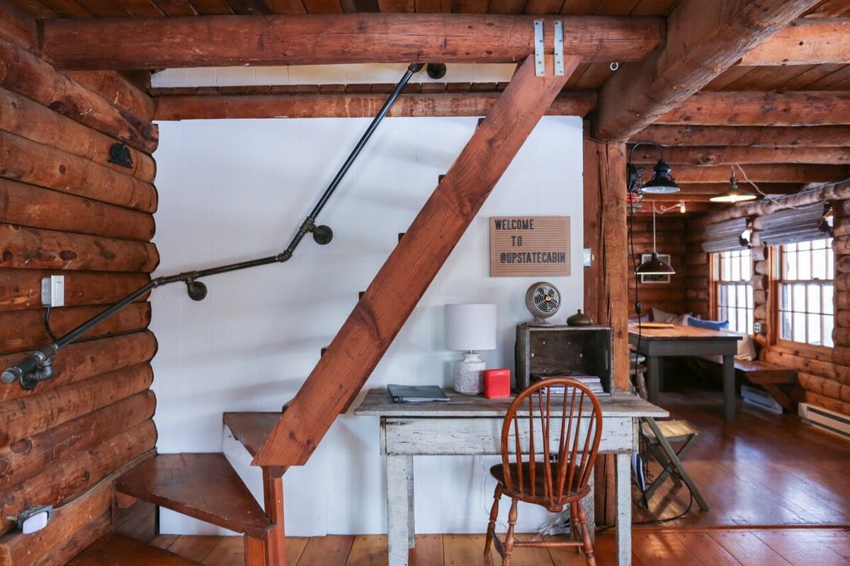 Inviting entryway or common area with a rustic wooden staircase featuring a black pipe railing, set against a white wall and exposed log walls. A vintage-style desk with a wooden chair and a 'WELCOME TO @UPSTATECABIN' sign creates a cozy workspace.