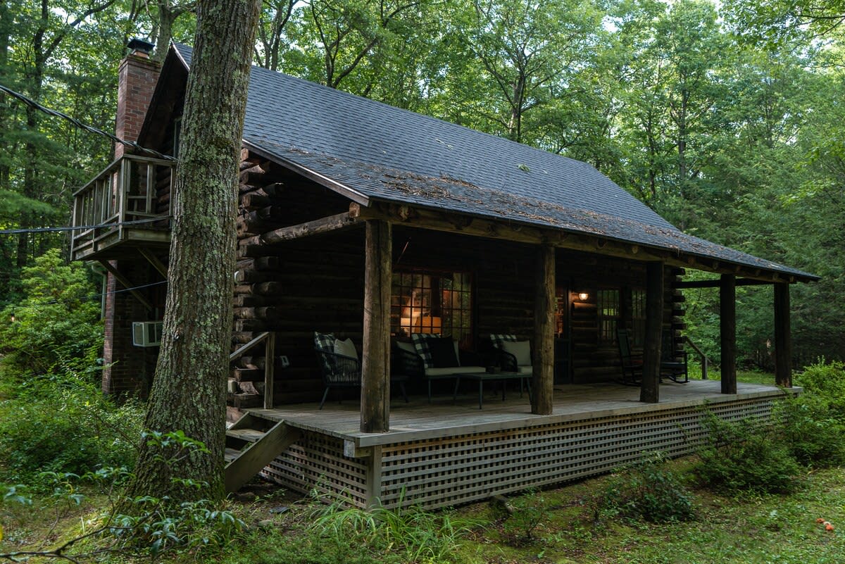 A rustic log cabin nestled in a lush green forest, featuring a dark shingle roof and a wide covered porch with outdoor seating. A stone chimney is visible on the left side, and a wooden deck extends from the front.