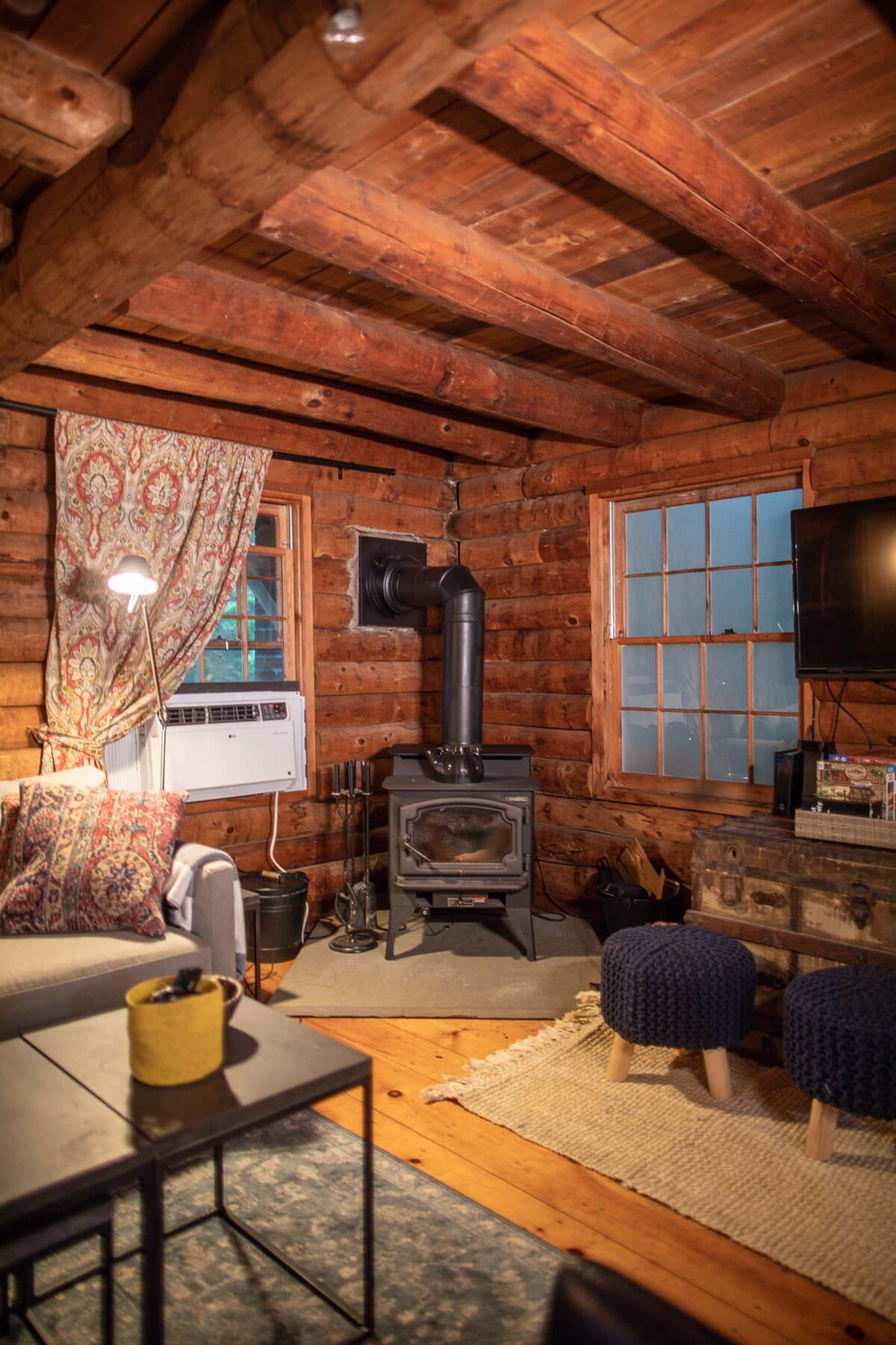 Close-up view of the rustic log cabin living room, highlighting the black wood-burning stove with a stovepipe extending through the log wall. A modern air conditioning unit is mounted above a patterned curtained window, with two blue poufs on a natural fiber rug.