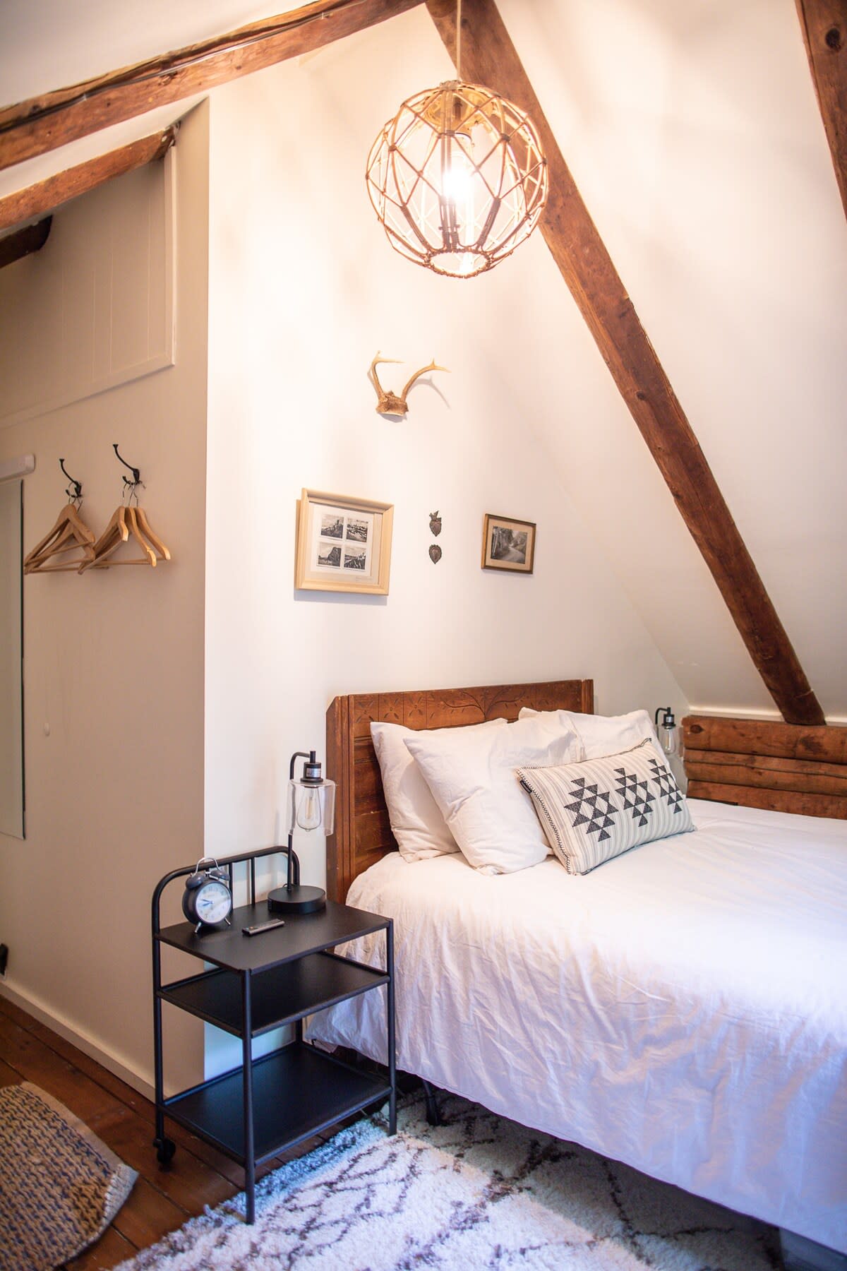 Cozy attic bedroom with a white vaulted ceiling and exposed wood beams, featuring a queen-sized bed with white bedding and a patterned throw pillow. A black metal nightstand with a lamp sits beside the bed, and a decorative antler mount adorns the wall.