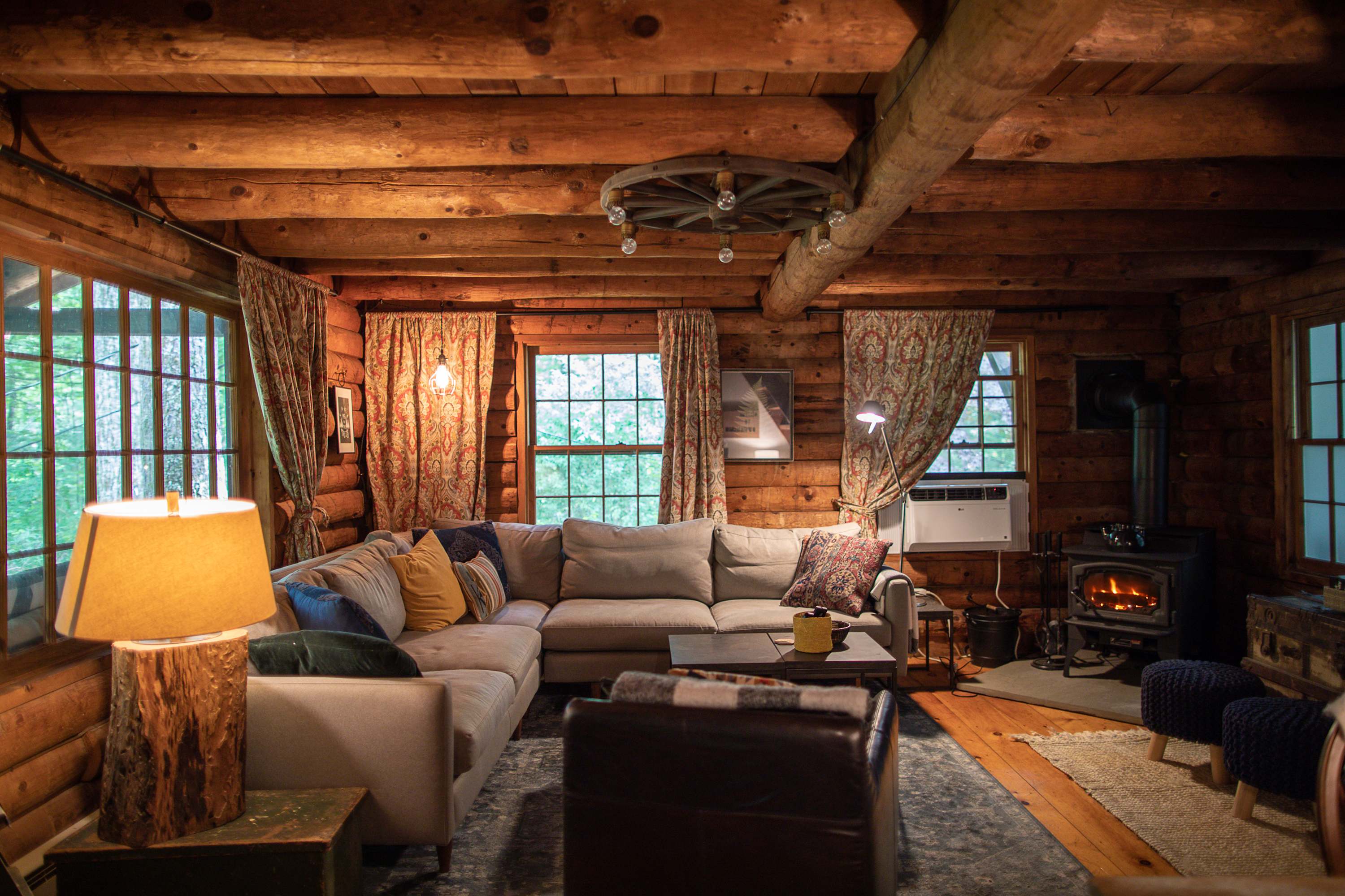 Cozy log cabin living room featuring a large L-shaped sectional sofa with throw pillows, a wood-burning stove, and rustic log walls and ceiling beams. Warm lighting from lamps and a wagon wheel chandelier creates an inviting atmosphere.