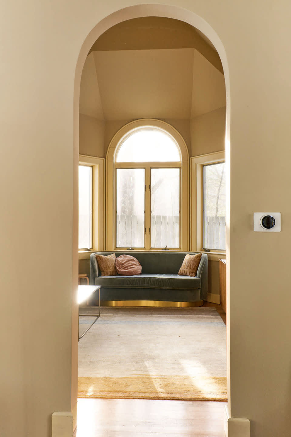 An elegant arched doorway opens to a cozy sitting nook featuring a curved green velvet sofa beneath a large bay window with an arched top. The room has light-colored walls and a patterned rug.