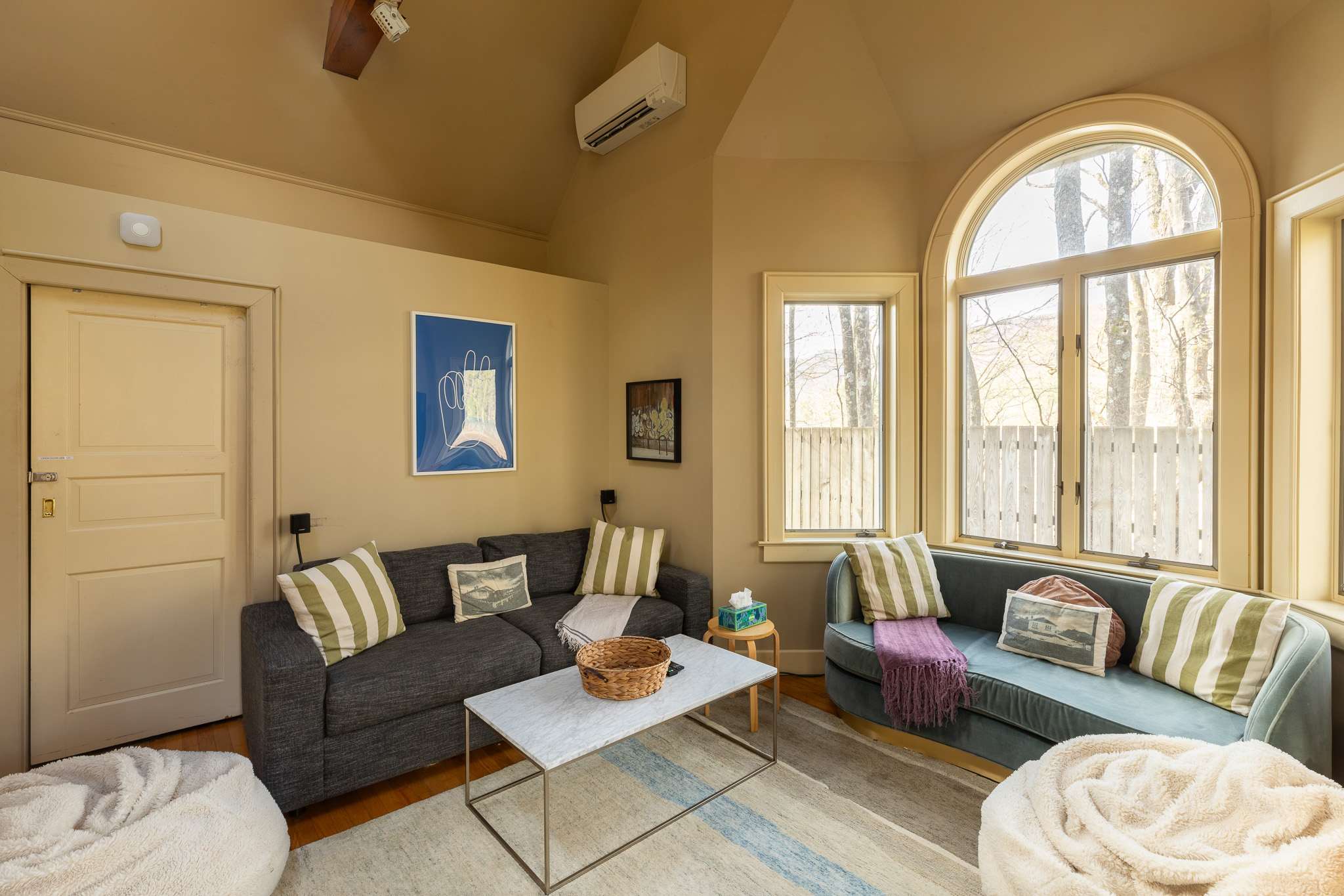 A secondary living area features a dark gray sofa and a curved teal velvet sofa, both adorned with pillows, a marble-top coffee table, and a wall-mounted air conditioning unit, with natural light from an arched window.