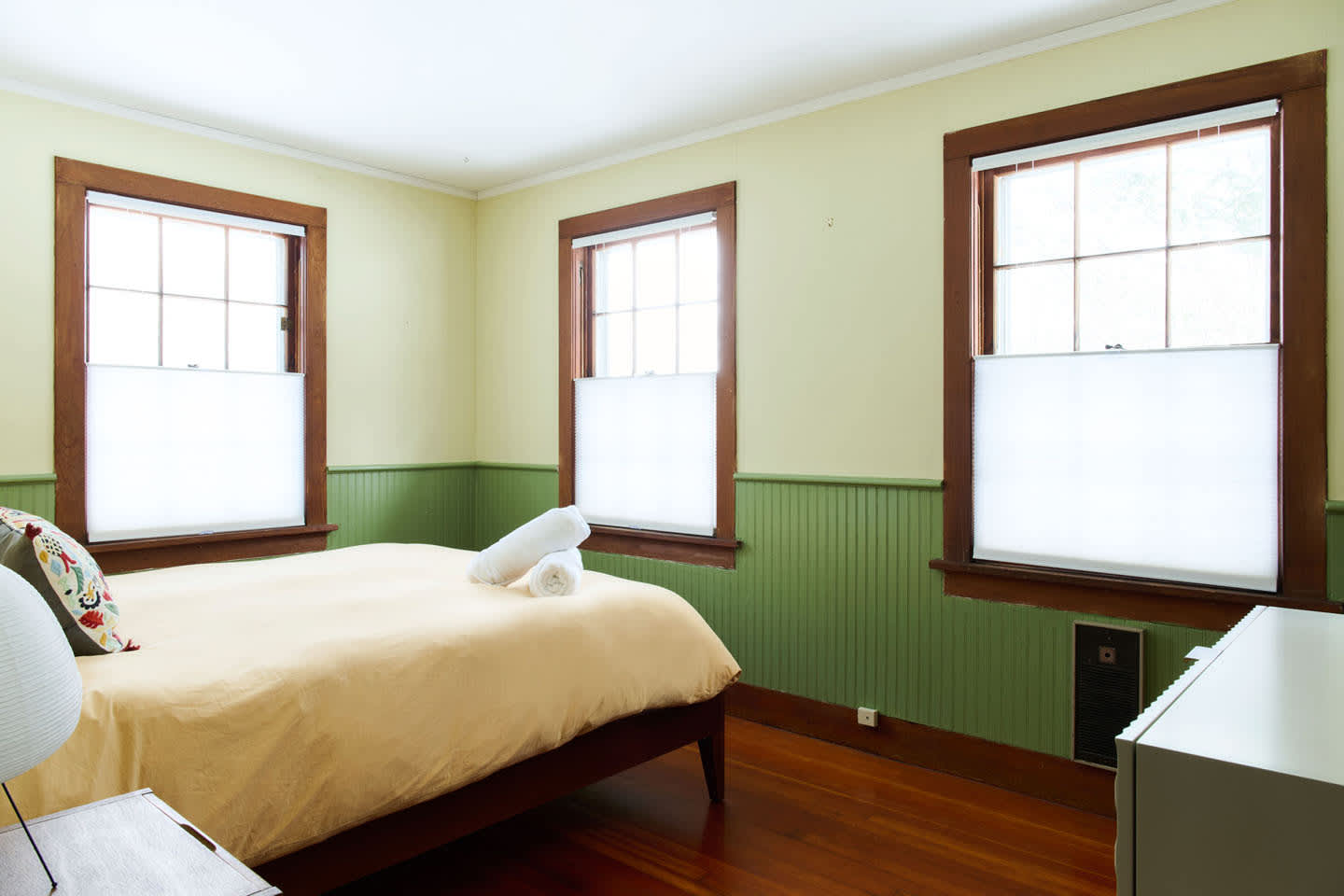A bright bedroom features light green walls with wainscoting, dark wood window frames, a bed with yellow bedding and white towels, and light-filtering blinds on three windows.