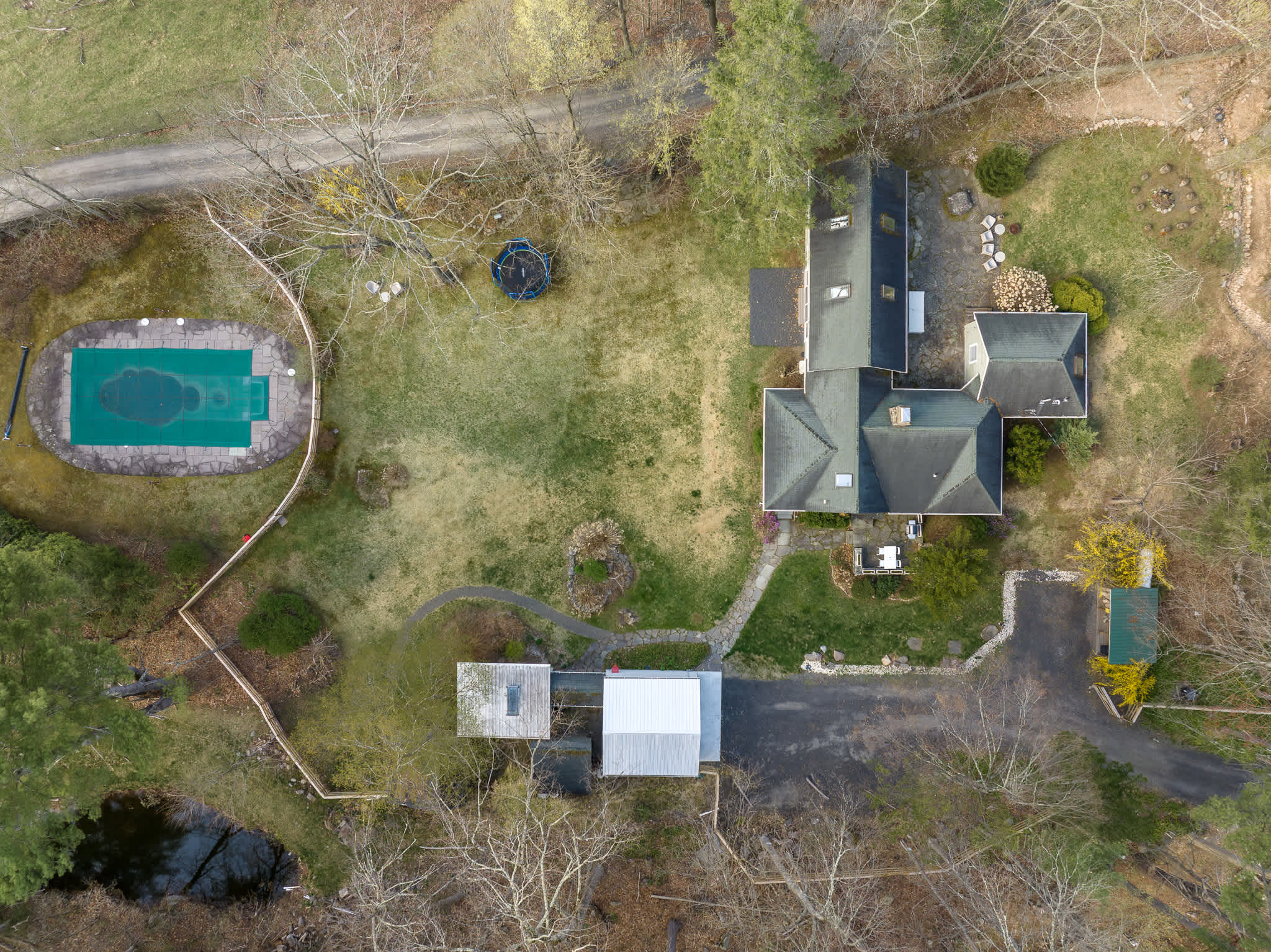 Overhead aerial view of the property, revealing a green-sided house, a large grassy yard, and a rectangular swimming pool covered with a blue tarp. The property is surrounded by dense woodland.