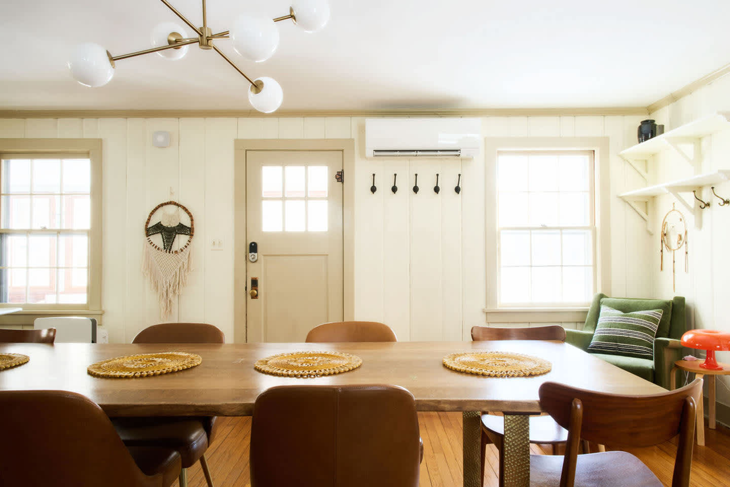 A dining area with a long wooden table and multiple chairs is illuminated by a modern Sputnik-style chandelier. White vertical paneling covers the walls, and a door with a window leads outside.