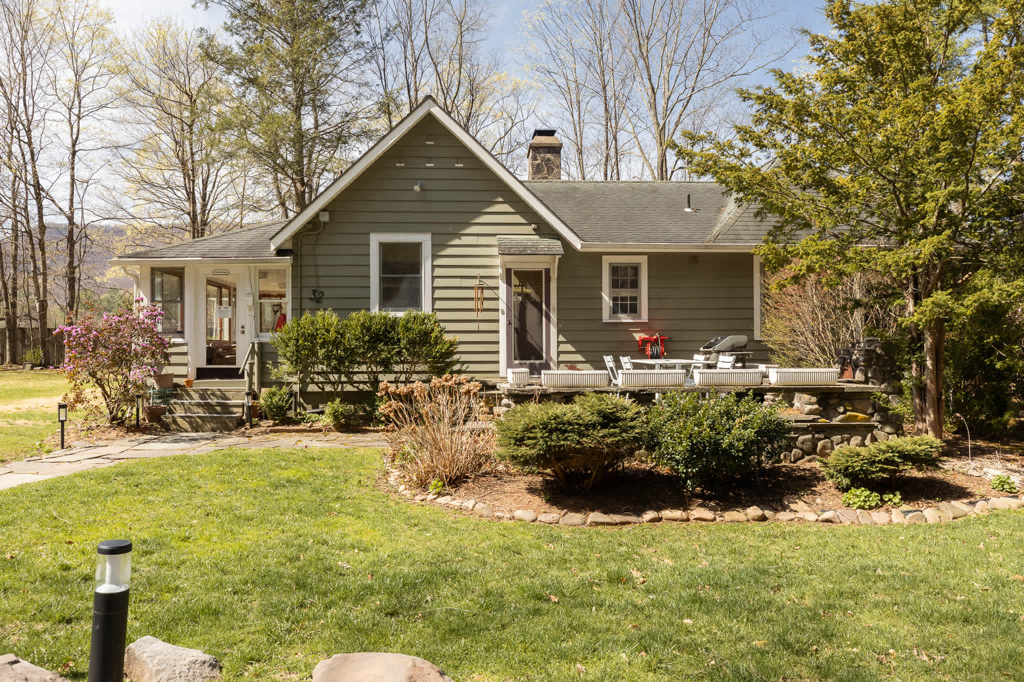 Front exterior of a single-story house with green siding, a dark front door, and a small stone patio with white outdoor chairs and a grill. Lush green lawn and mature trees surround the property.