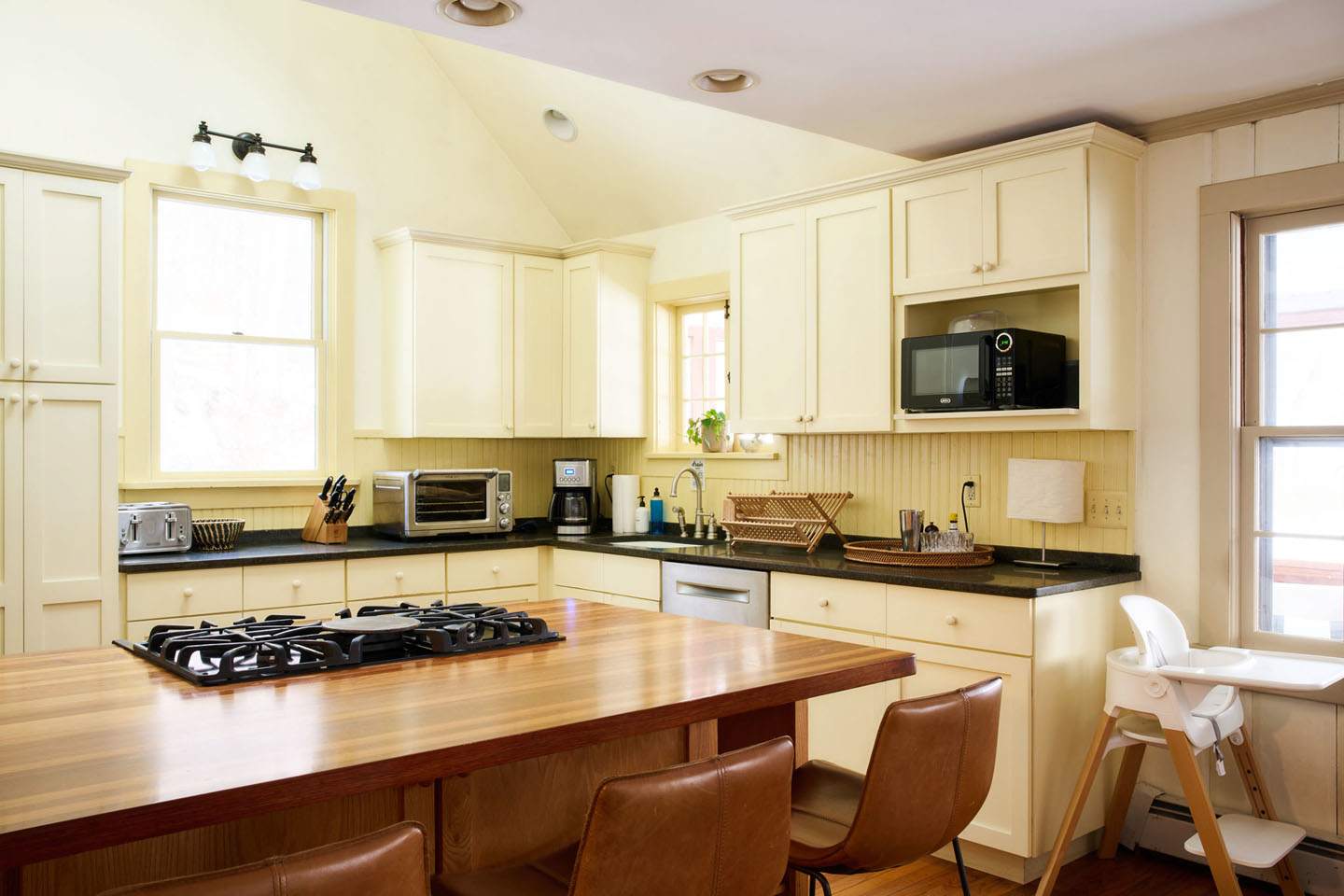 A spacious kitchen with light yellow cabinets, dark countertops, and a large wooden island featuring a gas range. Appliances include a microwave, toaster oven, and coffee maker, with hardwood floors throughout.