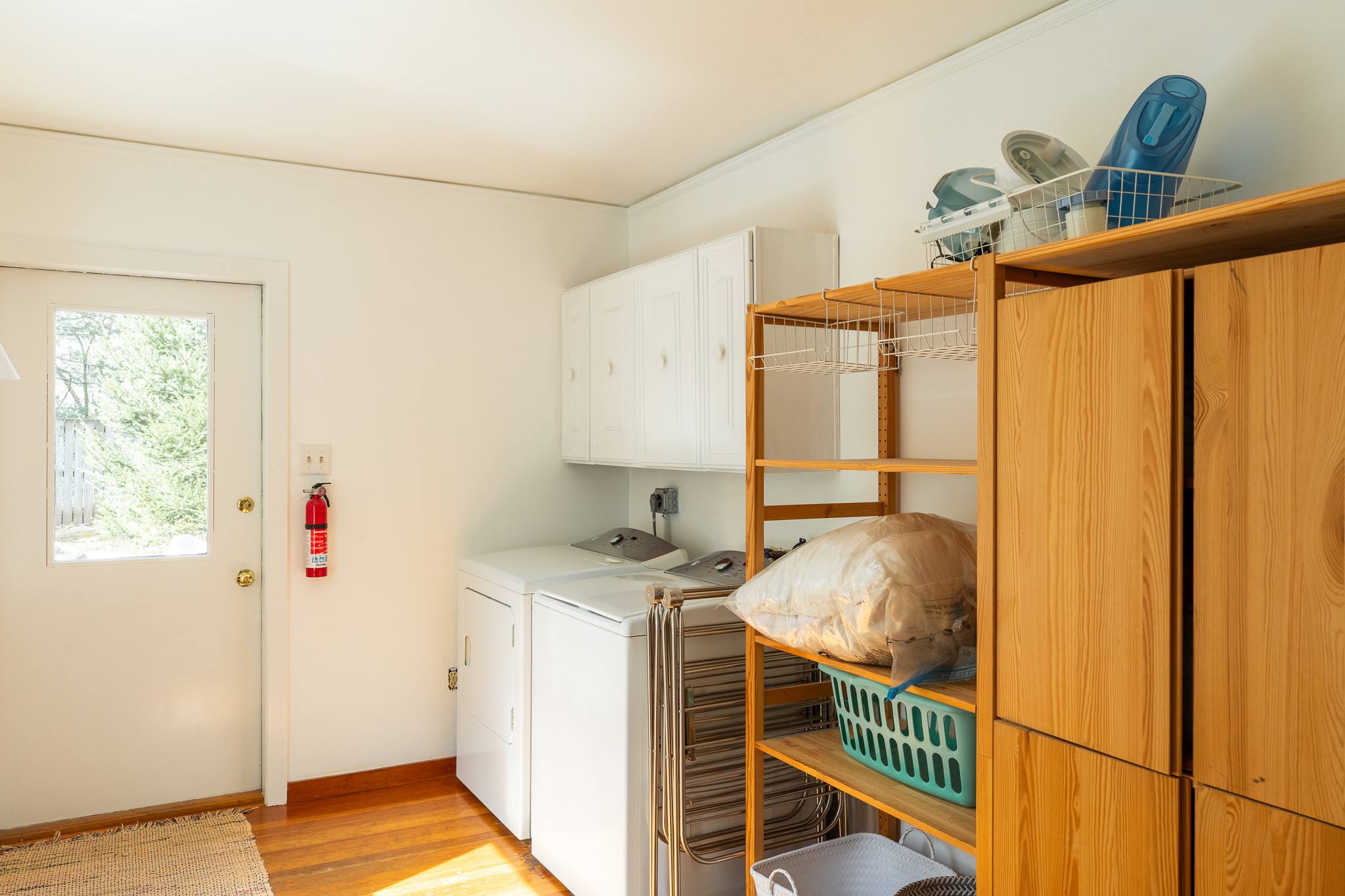 A functional laundry room equipped with white washer and dryer units, white upper cabinets, and open wooden shelving stocked with supplies. A glass-paned door provides access to the outdoors.