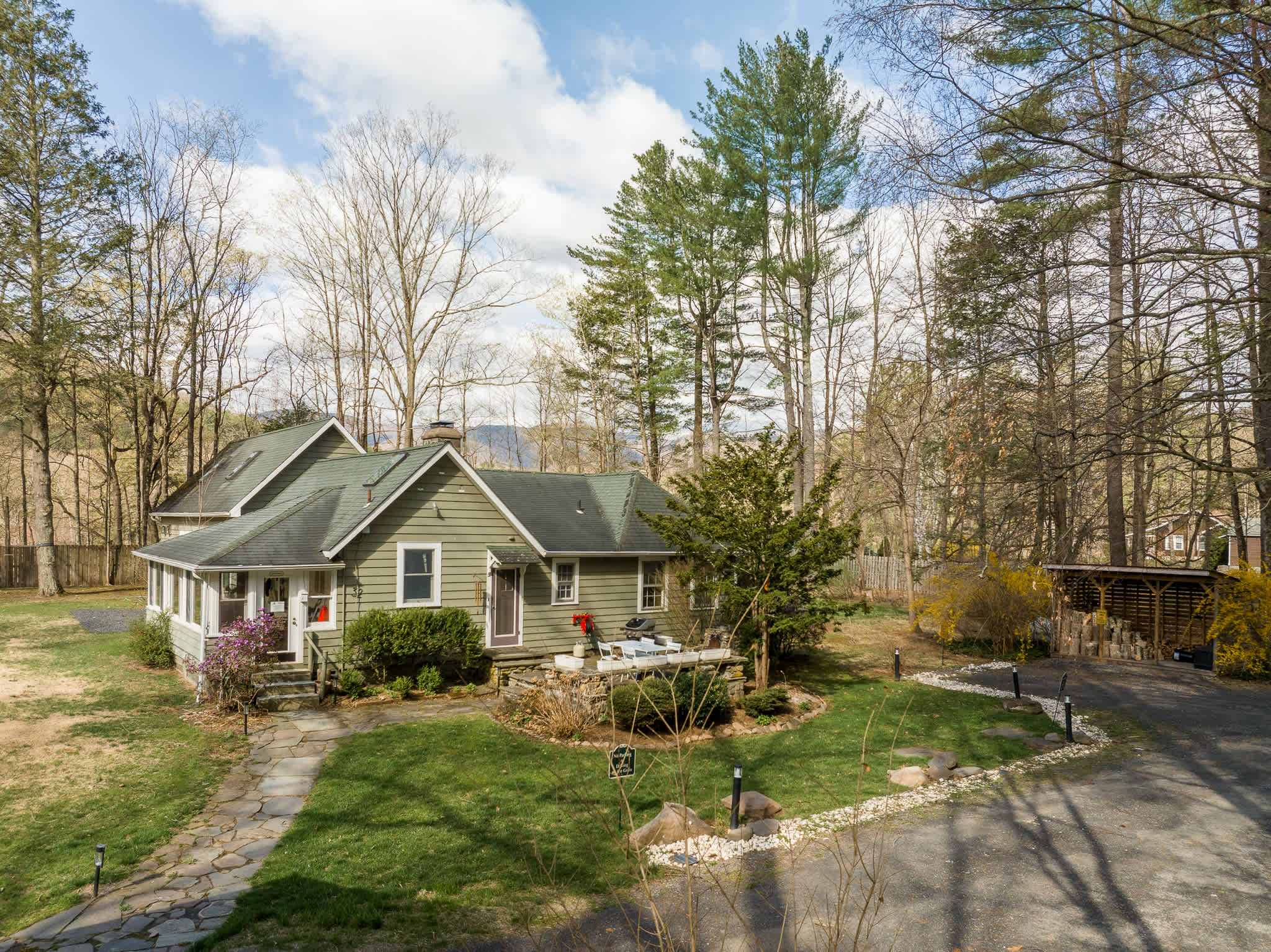 Elevated view of the green-sided house with a stone pathway leading to the front door, surrounded by a manicured lawn and mature trees. A gravel driveway is visible on the right, with distant mountains in the background.