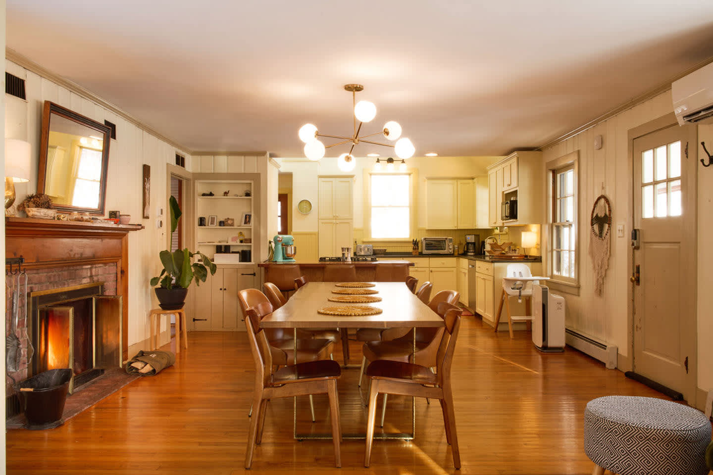 A wide view of the open-concept dining and kitchen area showcases a long wooden dining table with chairs, a brick fireplace, and a yellow-toned kitchen in the background. Hardwood floors extend throughout the space.