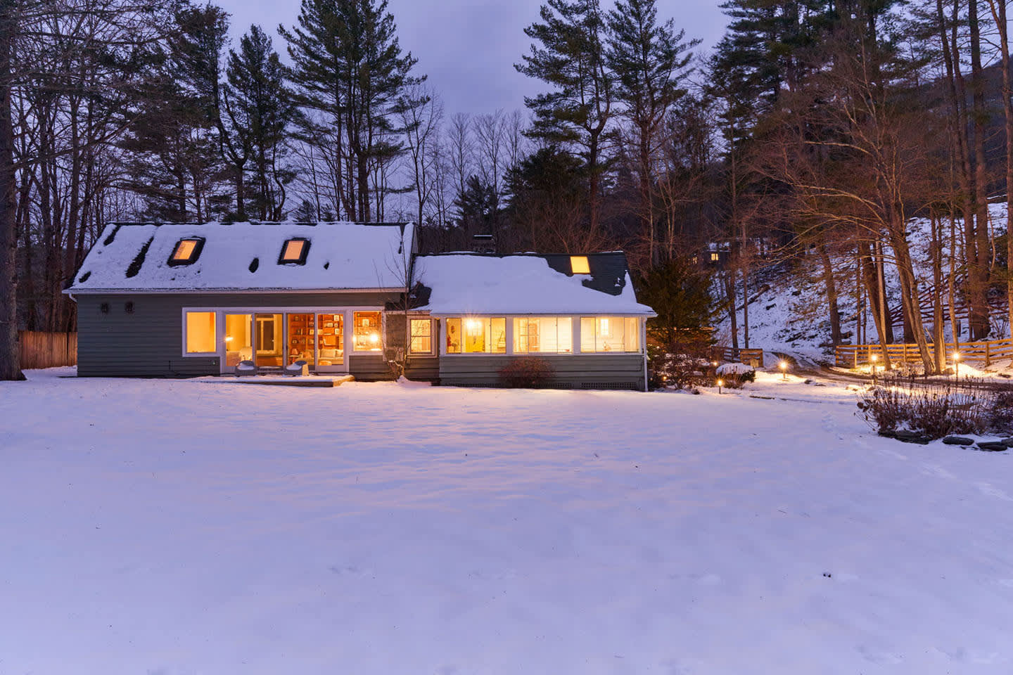 The back exterior of the house is captured at dusk, blanketed in snow, with warm interior lights illuminating the windows. The multi-sectioned house is nestled among tall, dark trees.