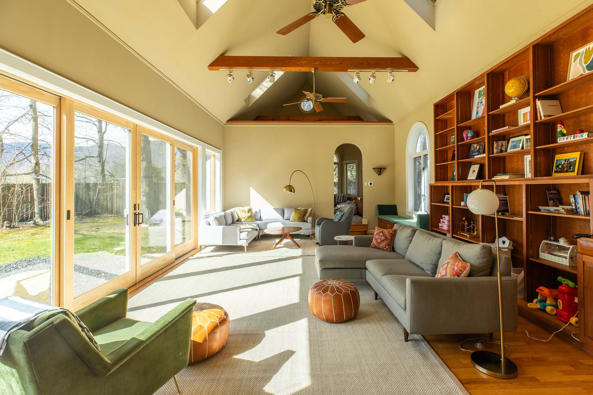 A sunlit living room boasts vaulted ceilings, exposed wooden beams, and large sliding glass doors that open to an outdoor yard, featuring a gray sectional sofa, a light gray sofa, and a green armchair.