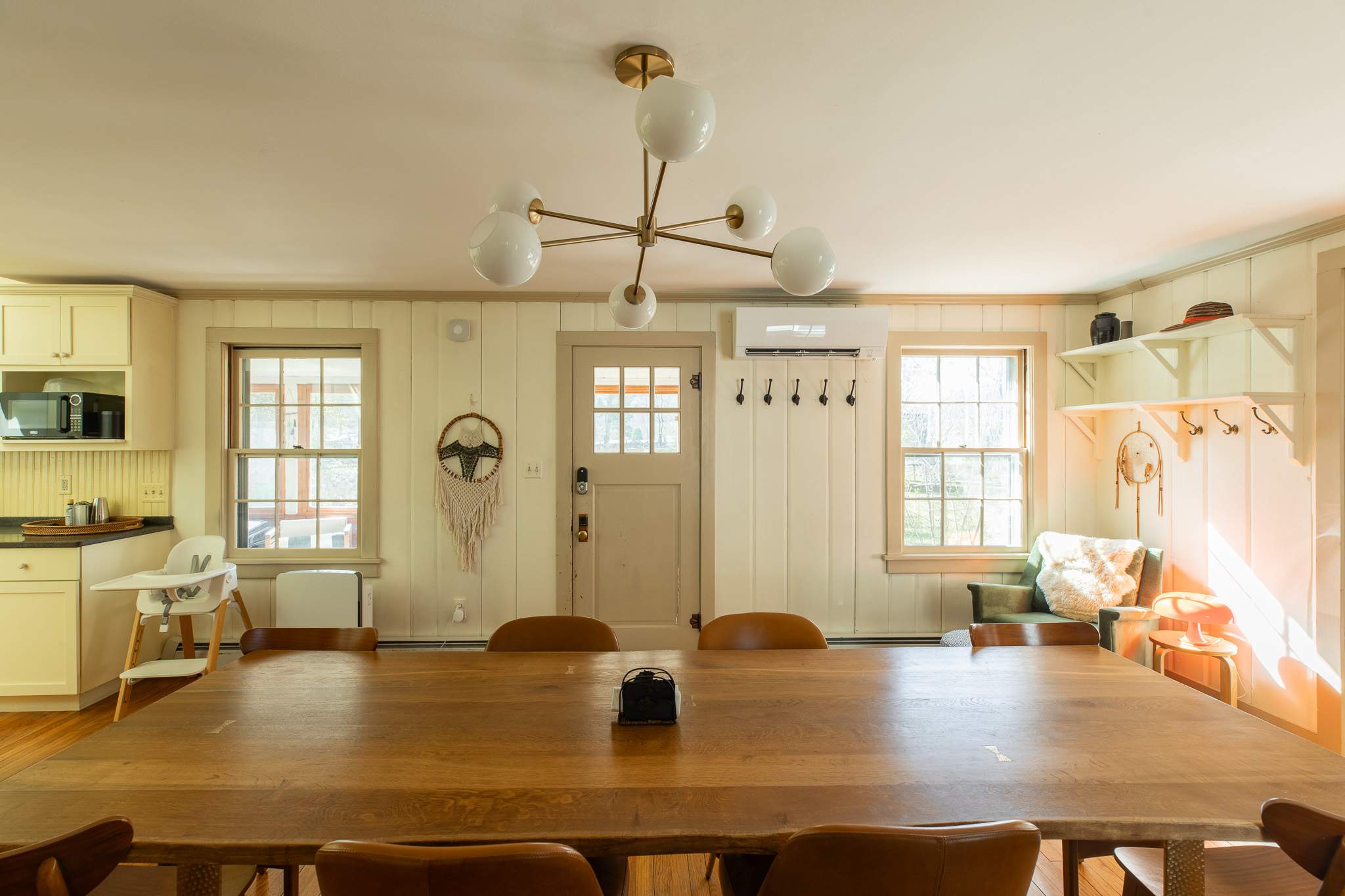A bright dining room features a long wooden table with leather chairs, a modern brass chandelier, and light-colored paneled walls. A high chair is visible near the kitchen counter, and a split AC unit is mounted above a door.
