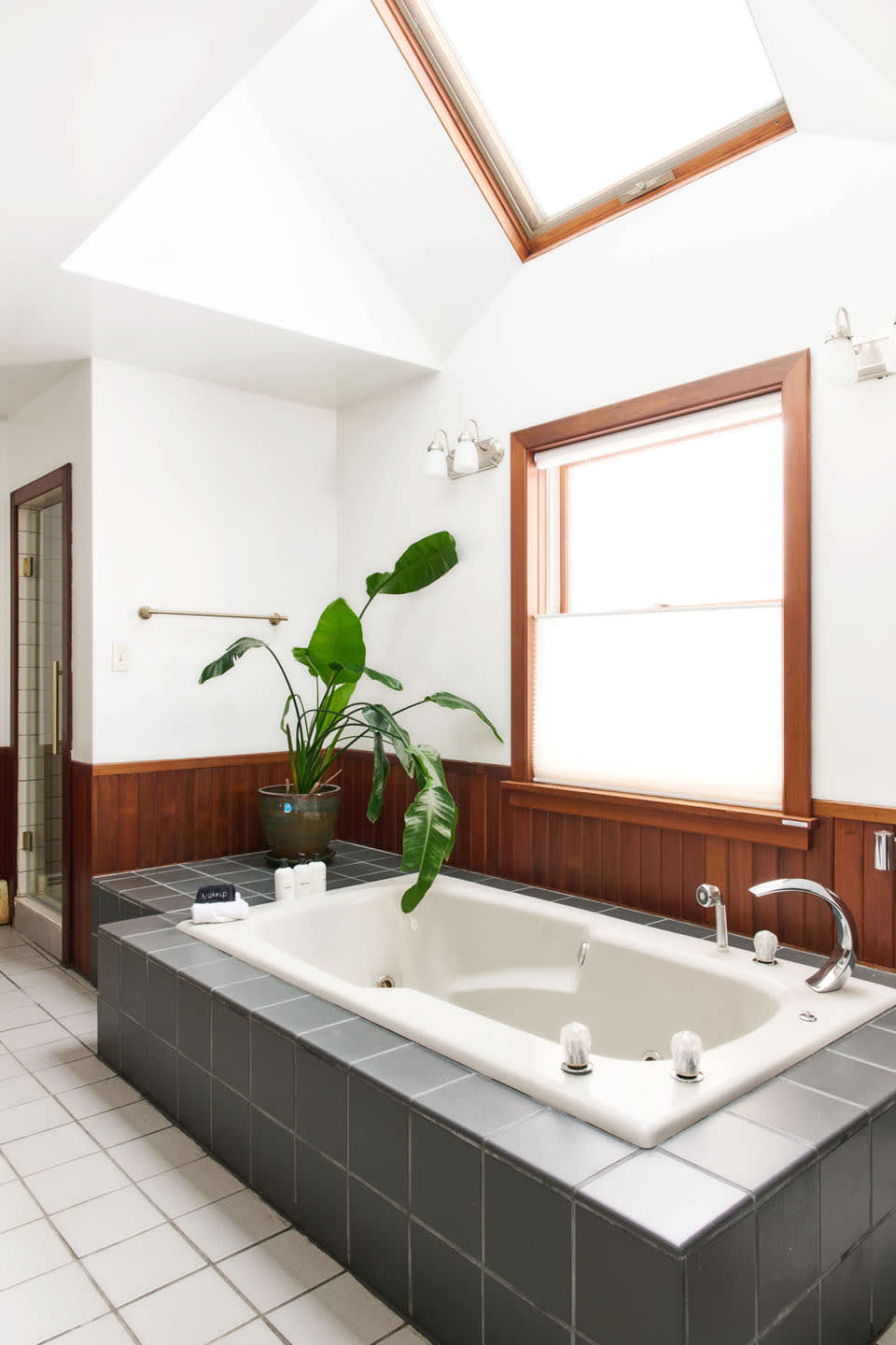 A bright bathroom features a large built-in jetted tub surrounded by dark gray tiles, illuminated by a skylight and a large window with warm wood trim. A tall green plant adds a touch of nature beside the tub.