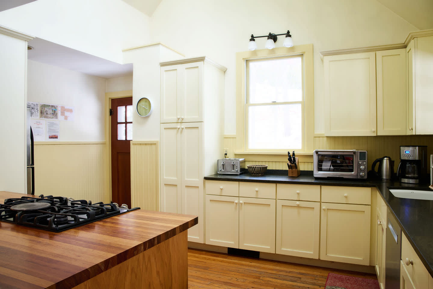 Corner view of the kitchen showcasing light yellow cabinets, dark countertops, and a wooden island with a gas range. A toaster and toaster oven are visible on the counter, with a tall pantry cabinet and a door leading to another room.