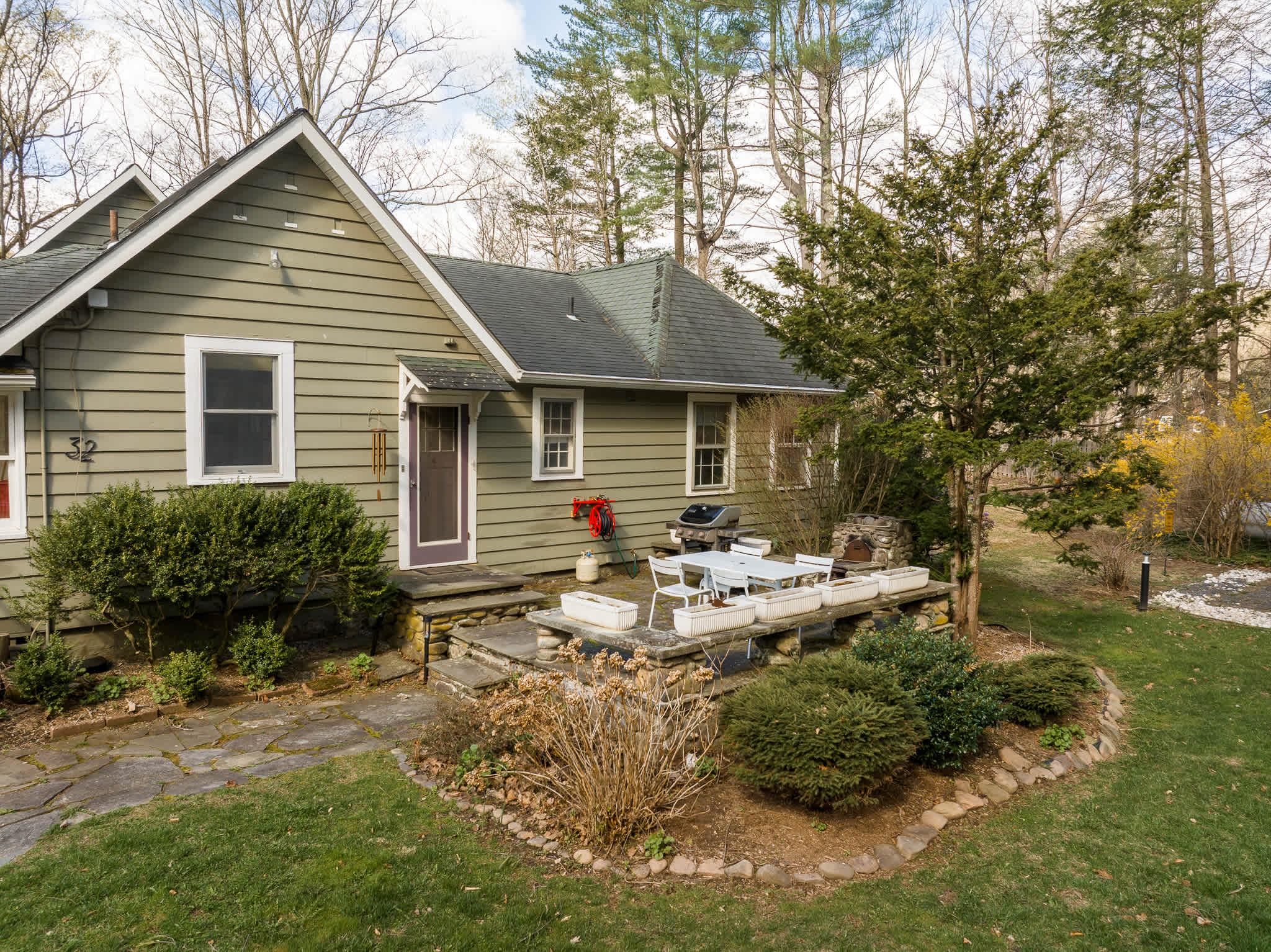 Elevated view of the front of the green-sided house, showcasing a stone patio with white outdoor furniture and a grill. Landscaped garden beds with shrubs and a small tree are in the foreground.