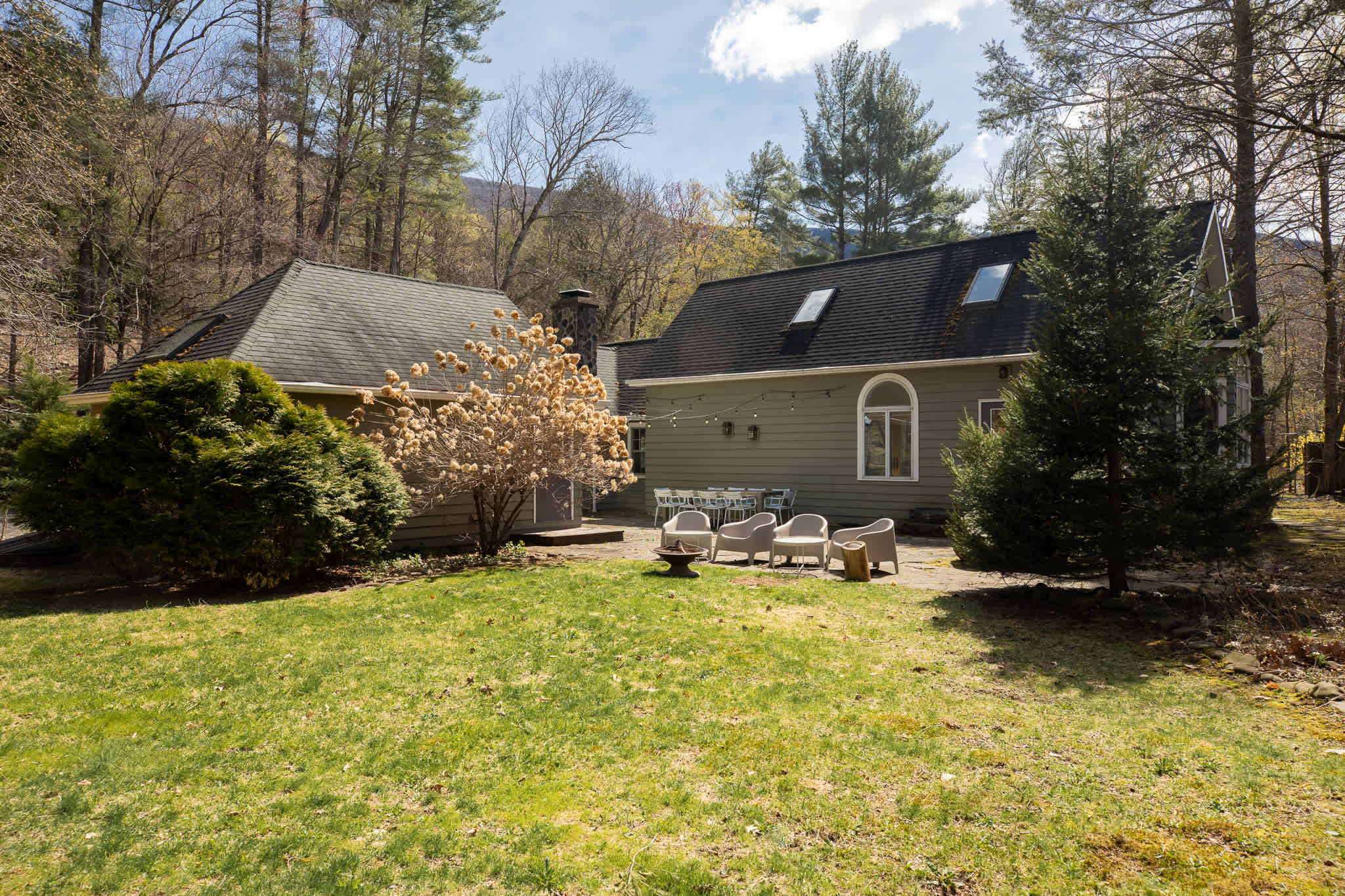 Rear exterior of the house featuring green siding, a large arched window, and a spacious flagstone patio with an outdoor dining table and separate lounge chairs. A large grassy yard extends towards a wooded area with distant mountains.