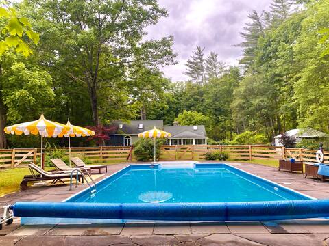 Outdoor swimming pool with bright blue water, surrounded by a stone deck with lounge chairs and two yellow and white striped umbrellas. The house is visible in the background, nestled among trees.