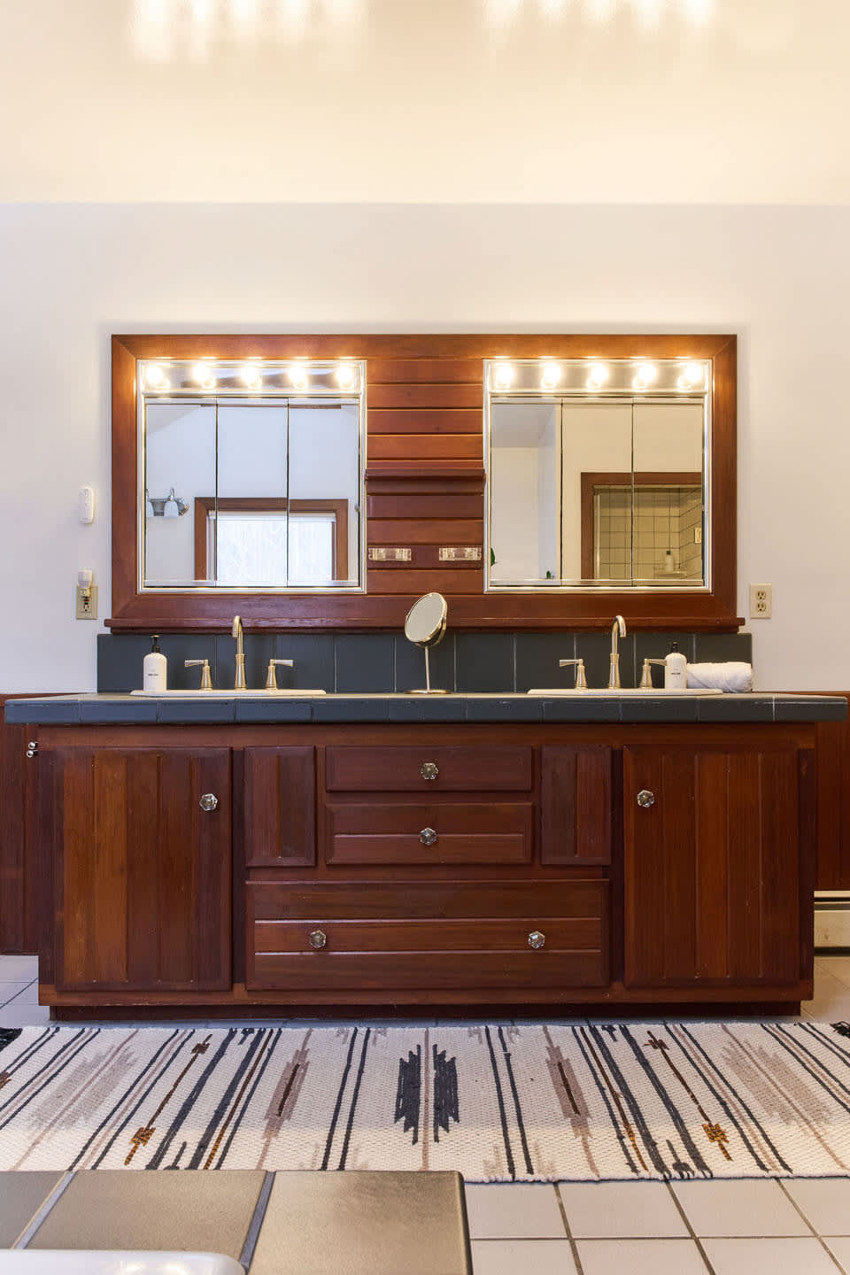 A rustic double vanity features dark wood cabinetry, two sinks with gold-toned faucets, and a dark tiled countertop. Two large mirrors with Hollywood-style strip lighting are mounted above, with a patterned rug on the floor.
