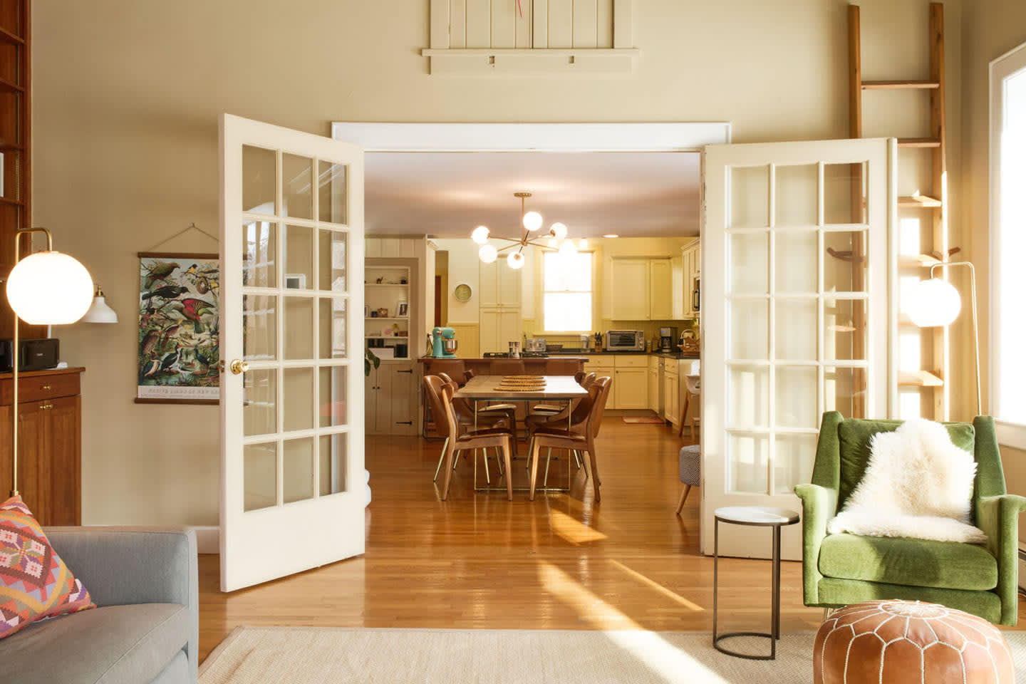 A spacious living room with light walls and hardwood floors features a gray sofa, a green armchair with a faux fur throw, and a round ottoman. French doors open into a dining area and kitchen beyond.