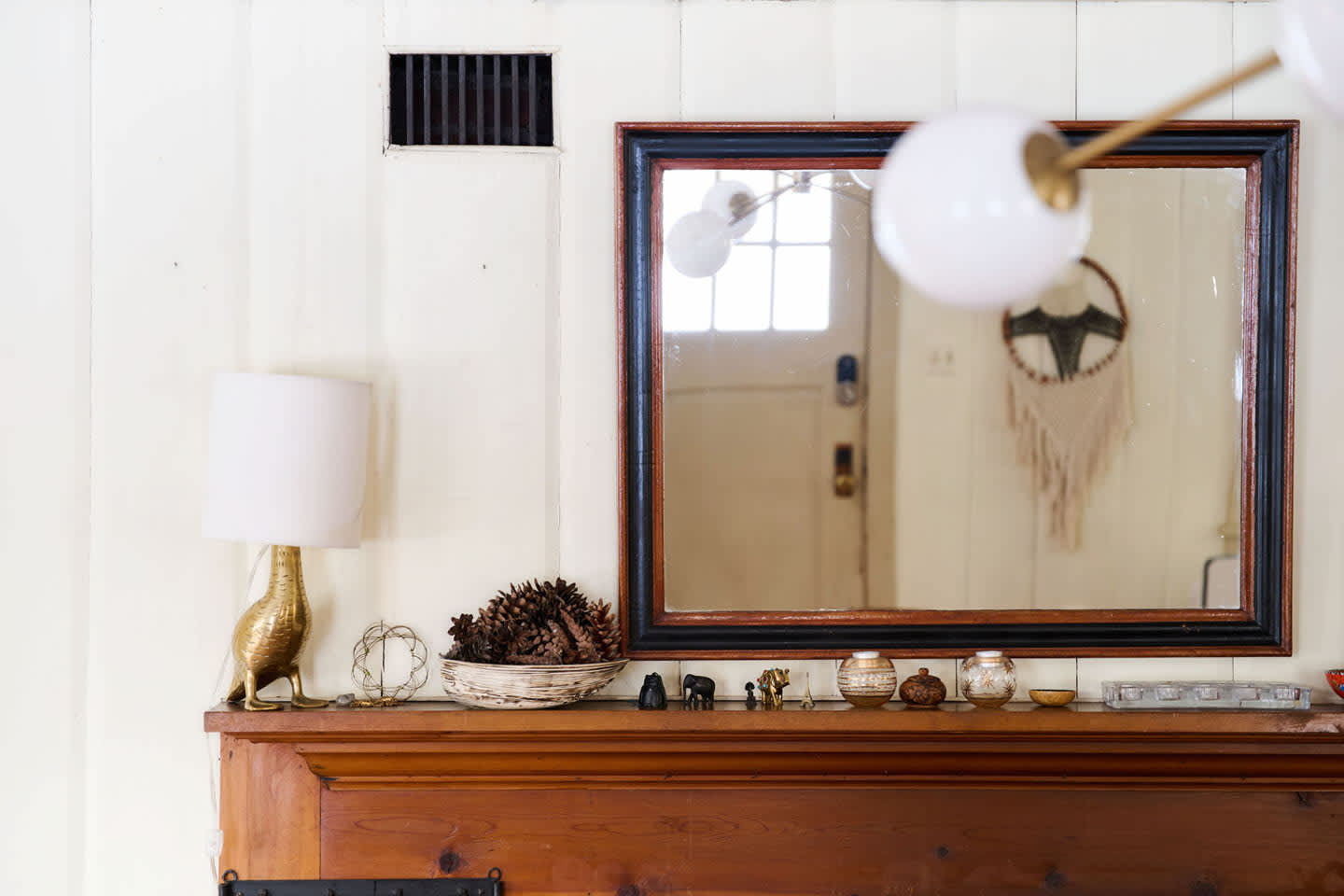 A wooden mantelpiece adorned with decorative items including a gold lamp, pinecones in a basket, and small animal figurines. A large mirror above reflects the dining room and a macrame wall hanging.