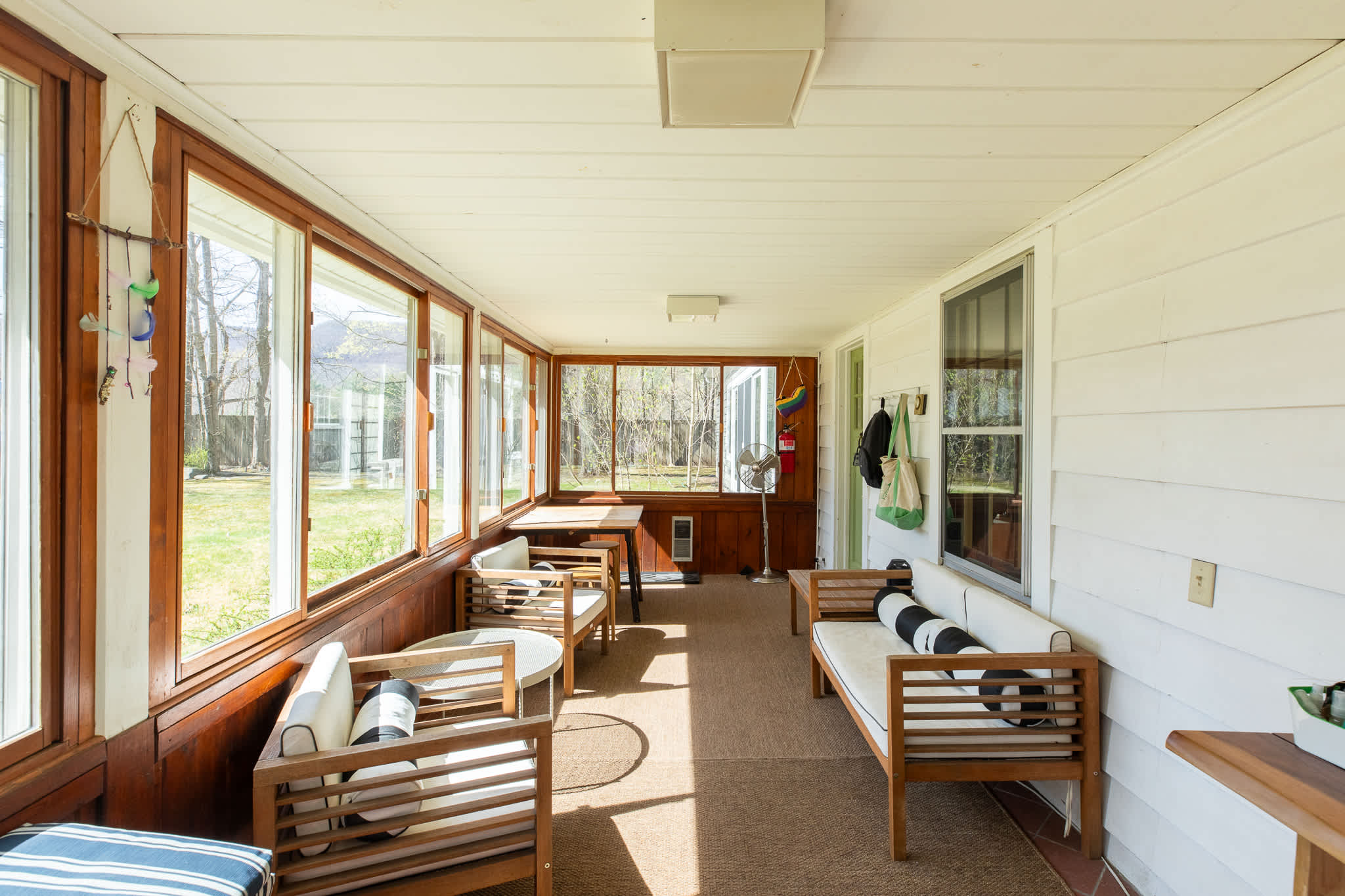 An enclosed porch or sunroom is filled with natural light from multiple large windows offering views of the outdoors. It is furnished with light wood-framed seating, striped cushions, a small table, and a ceiling fan.