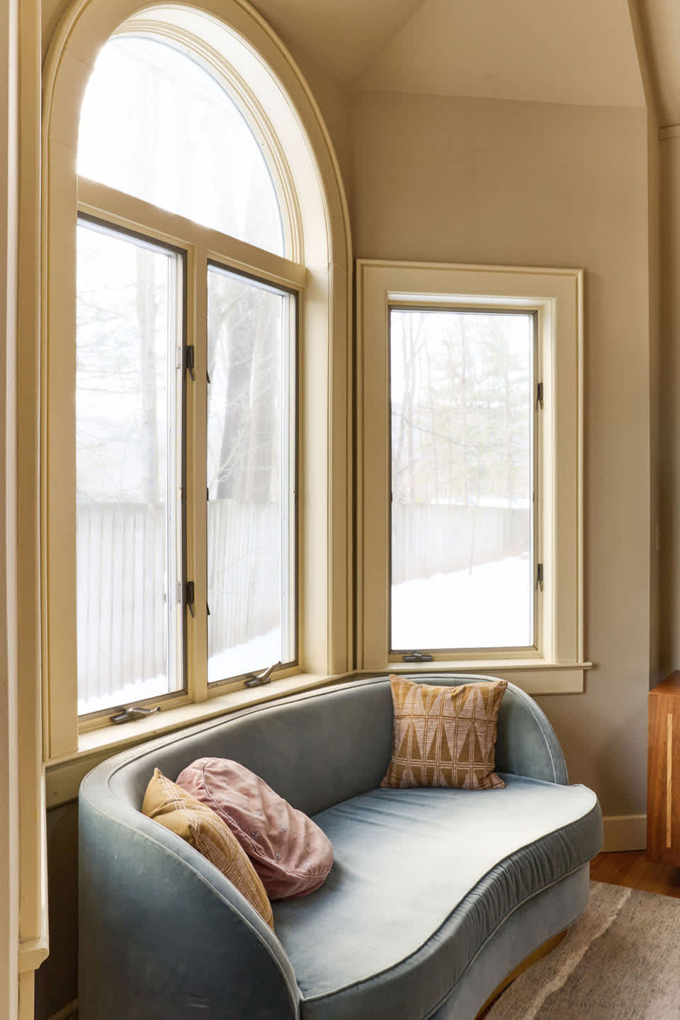 A cozy reading nook features a light blue velvet curved sofa with decorative pillows, positioned beneath a large arched window and a rectangular window, offering a bright view of a snowy outdoor scene.