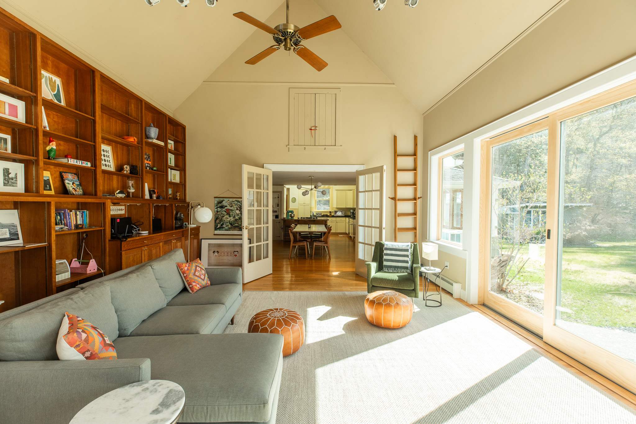 A spacious living room features a vaulted ceiling with a ceiling fan, a large grey sectional sofa, and extensive built-in wooden bookshelves. Natural light streams in through large glass doors leading to the yard.