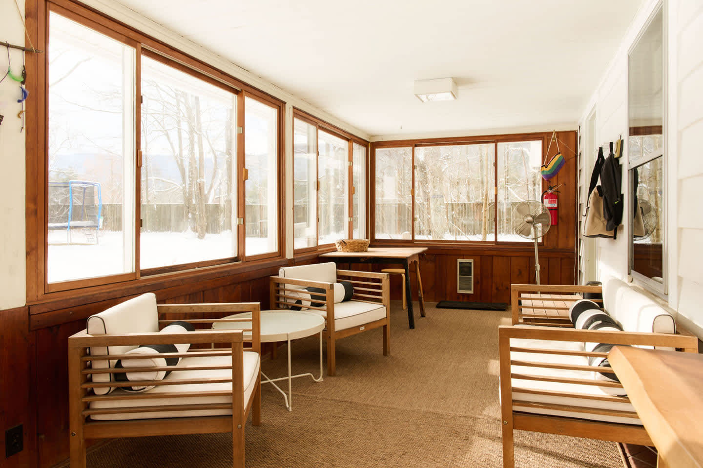 An enclosed sunroom with large windows offers snowy outdoor views, furnished with light wood chairs featuring white cushions and a small round table. Dark wood paneling lines the lower walls, and a ceiling fan is visible.