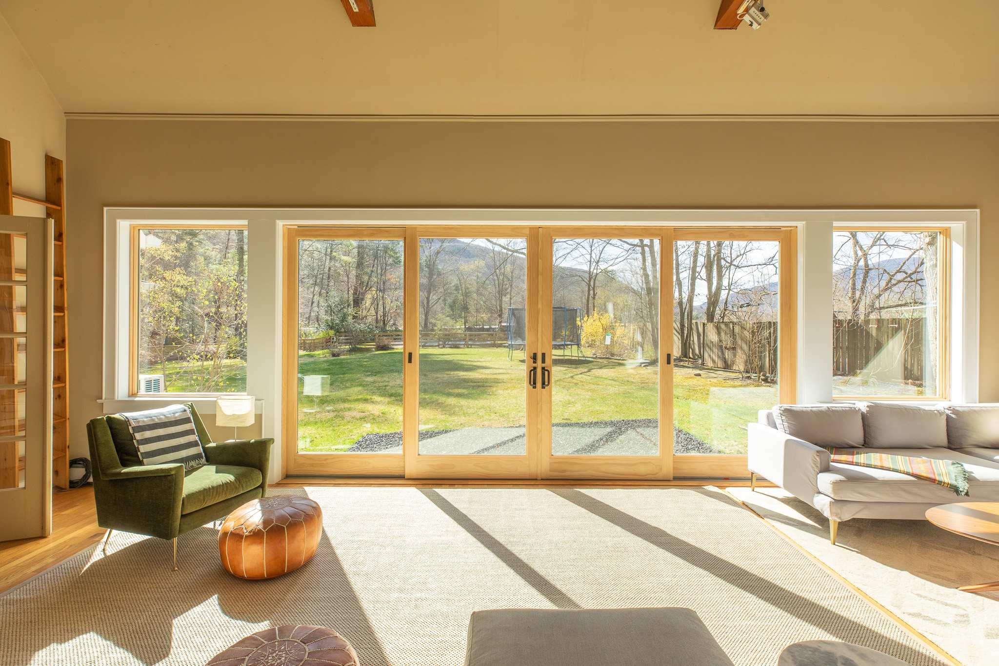 Large wooden-framed sliding glass doors offer a panoramic view of a lush green backyard with a trampoline and distant mountains, flanked by a green armchair and a light gray sofa.