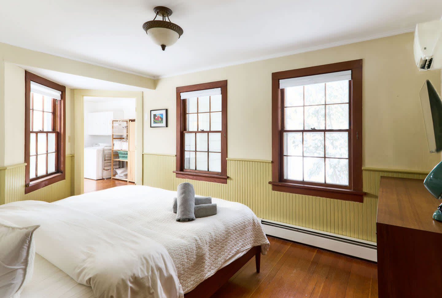 A spacious bedroom with light yellow walls and green wainscoting, featuring two large windows with dark wood trim. A white bed with grey towels is centered, and an open doorway leads to a laundry area.