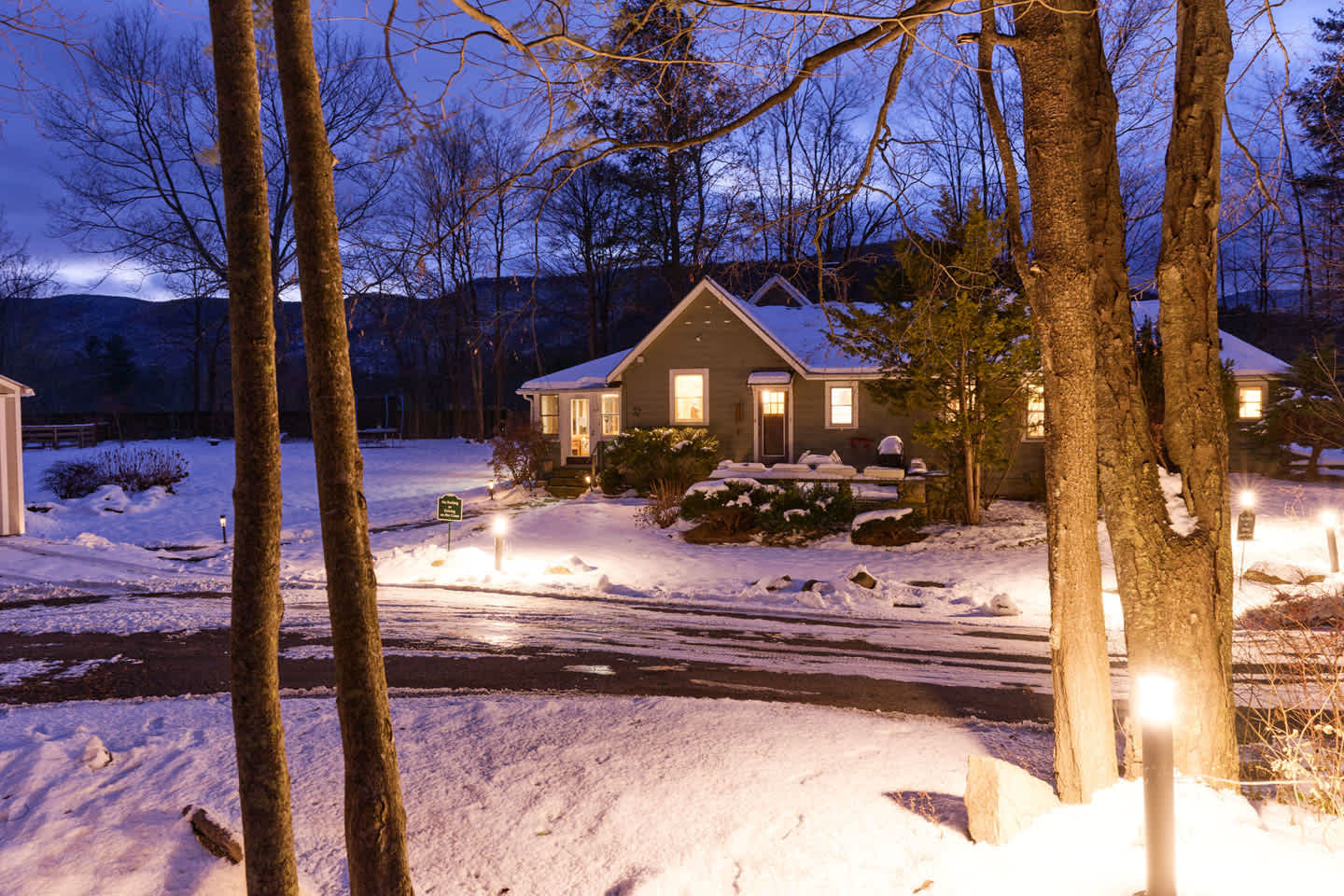 The front of the house is seen at dusk, covered in snow, with outdoor path lights illuminating the driveway and entrance. Bare trees frame the scene, and distant mountains are visible under the twilight sky.