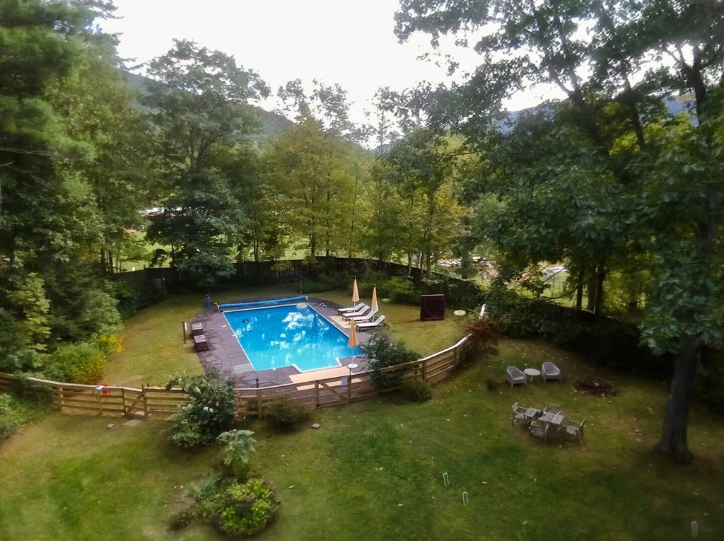 Aerial view of a large backyard featuring a rectangular blue swimming pool, surrounded by a wooden deck and fence. A grassy area with outdoor seating and a fire pit is also visible, all nestled within a lush, tree-filled landscape.