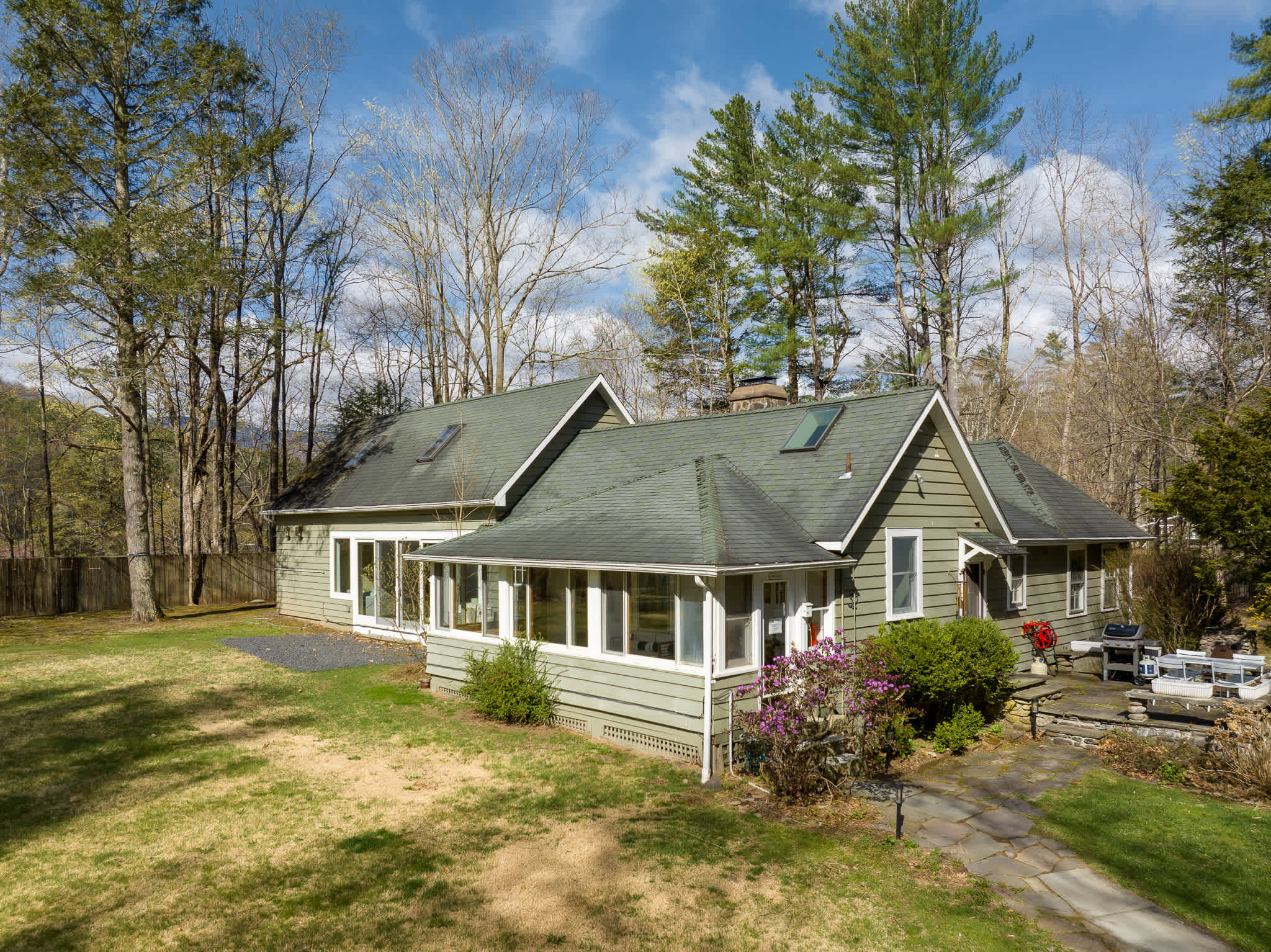 Side view of the green-sided house, featuring a sunroom or enclosed porch with multiple windows and a separate section with skylights on the roof. A large grassy area is in the foreground, surrounded by trees.