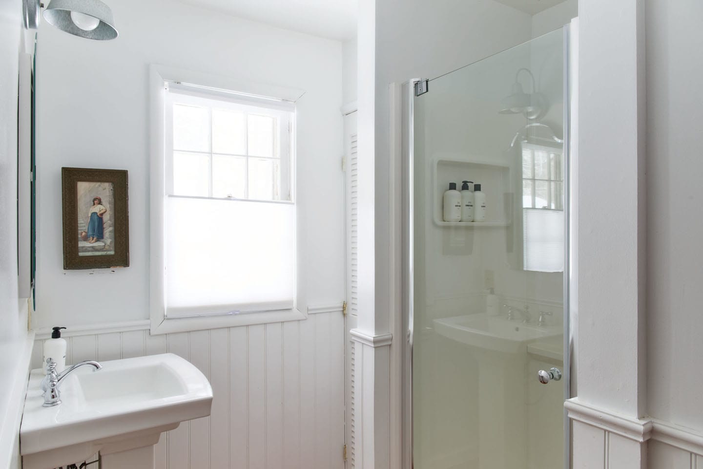 A clean white bathroom with beadboard wainscoting, a pedestal sink, and a glass-enclosed shower. A window with a privacy blind and a framed picture are visible on the wall.