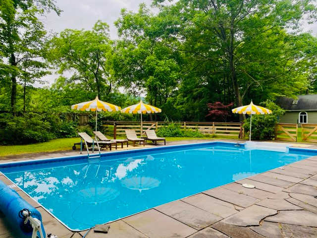 A rectangular blue swimming pool with stone coping is surrounded by a stone patio, featuring three yellow and white striped umbrellas and lounge chairs. Lush green trees and a wooden fence enclose the private outdoor space.
