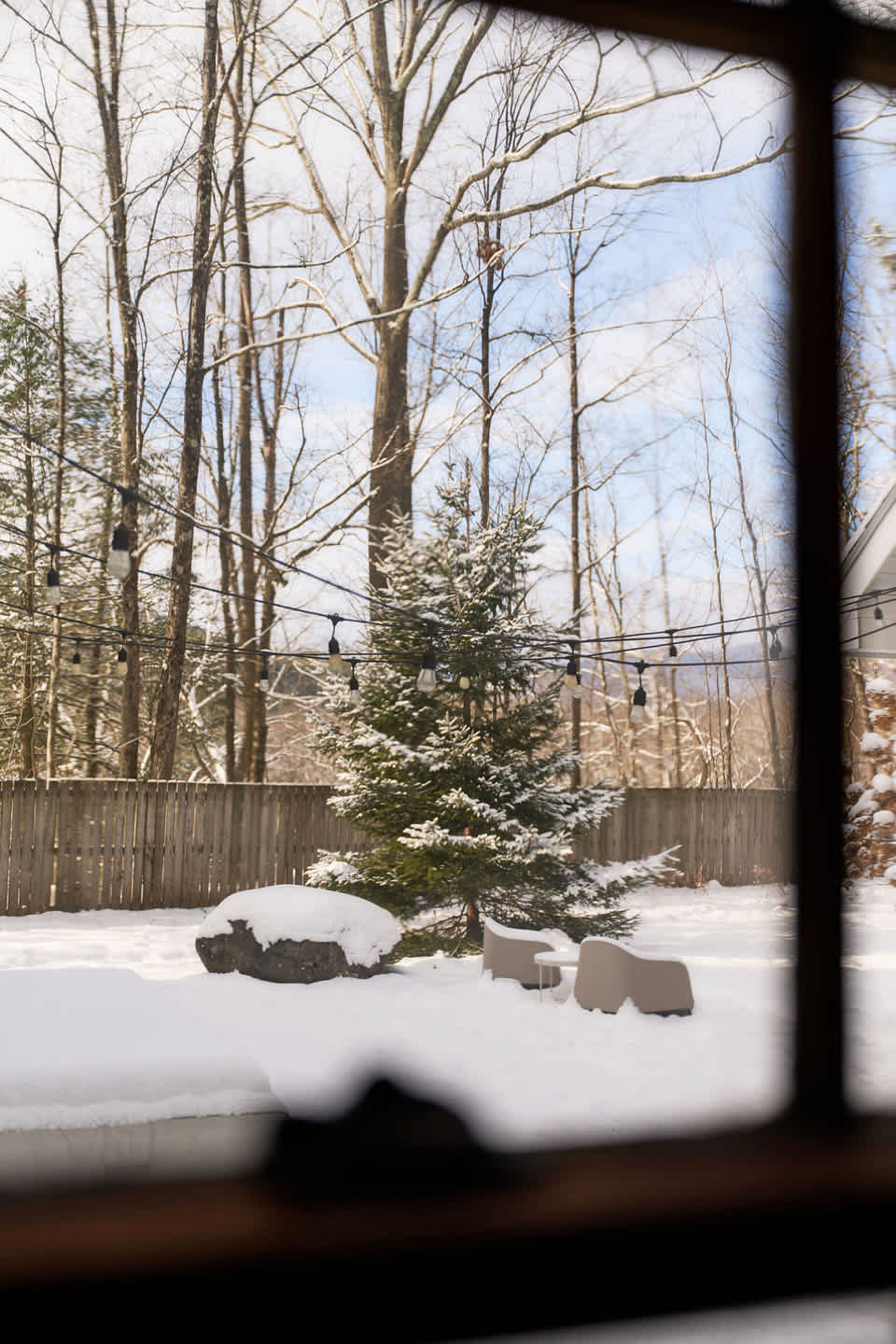 A view through a window reveals a snow-covered backyard featuring a small evergreen tree adorned with string lights, modern outdoor seating, and a large rock. Tall bare trees and a wooden fence complete the winter landscape.