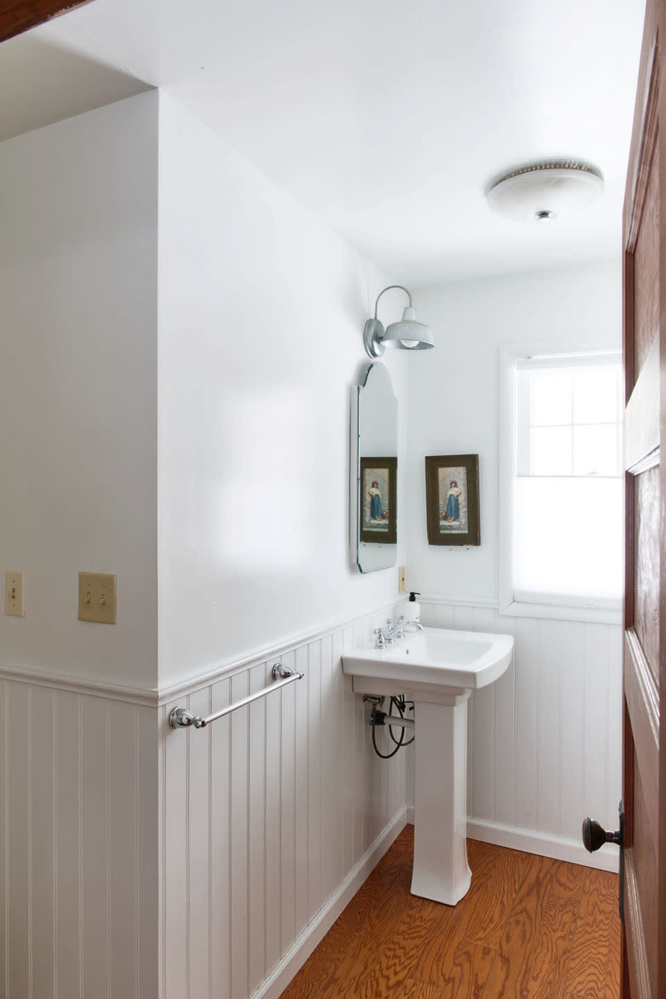 A bright white bathroom featuring beadboard wainscoting, a white pedestal sink with a mirror above it, and a window with a privacy blind. The room has warm hardwood floors and a dark wooden door.