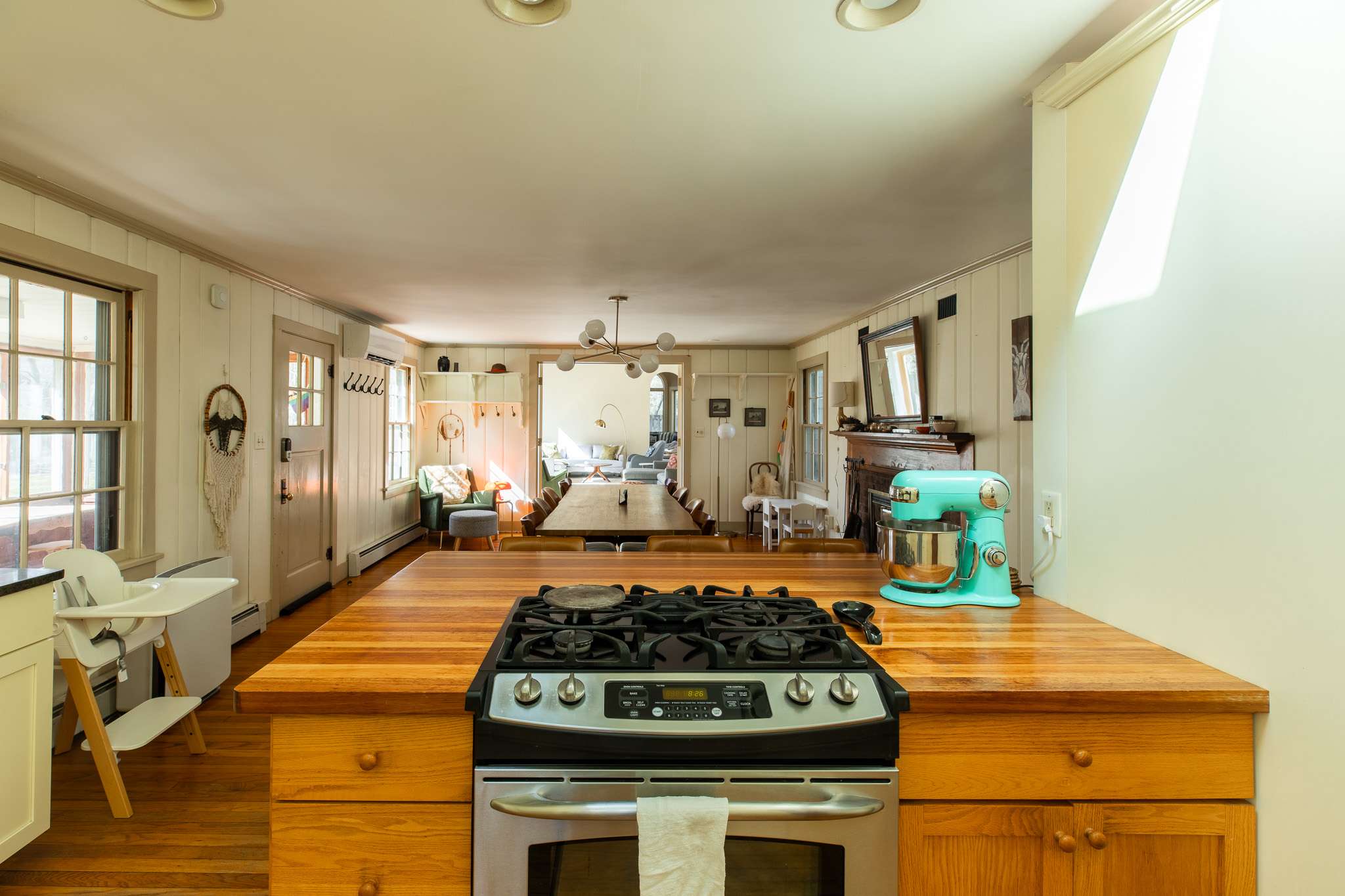 View from the kitchen island with a gas range, looking into an open-concept dining area with a long wooden table and a living room beyond. Light paneled walls and hardwood floors connect the spaces.