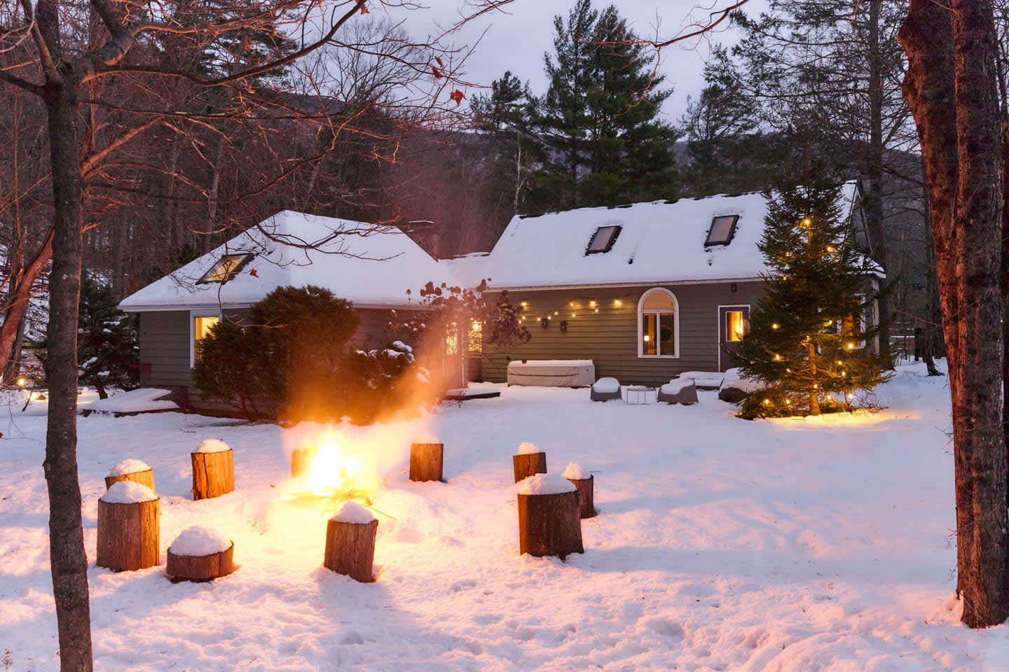 A glowing fire pit surrounded by tree stump seats illuminates a snow-covered backyard at dusk, with the house visible in the background adorned with festive lights. Bare trees frame the scene.