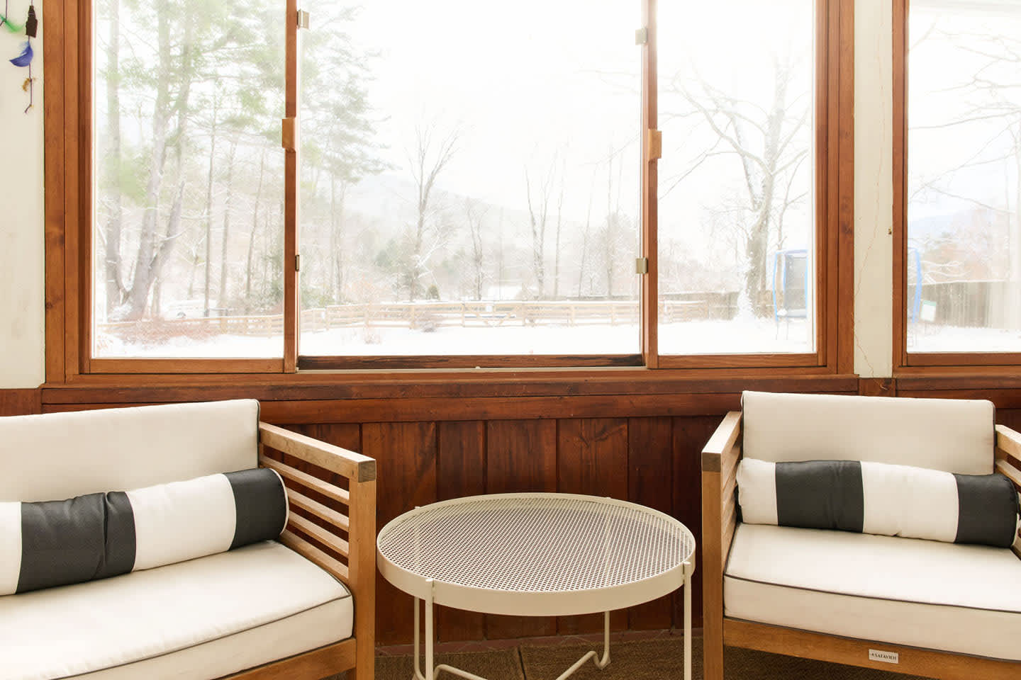 A close-up view of the enclosed sunroom highlights two light wood chairs with white cushions and black and white bolster pillows, flanking a small white round table. Large windows reveal a snowy, wooded landscape outside.