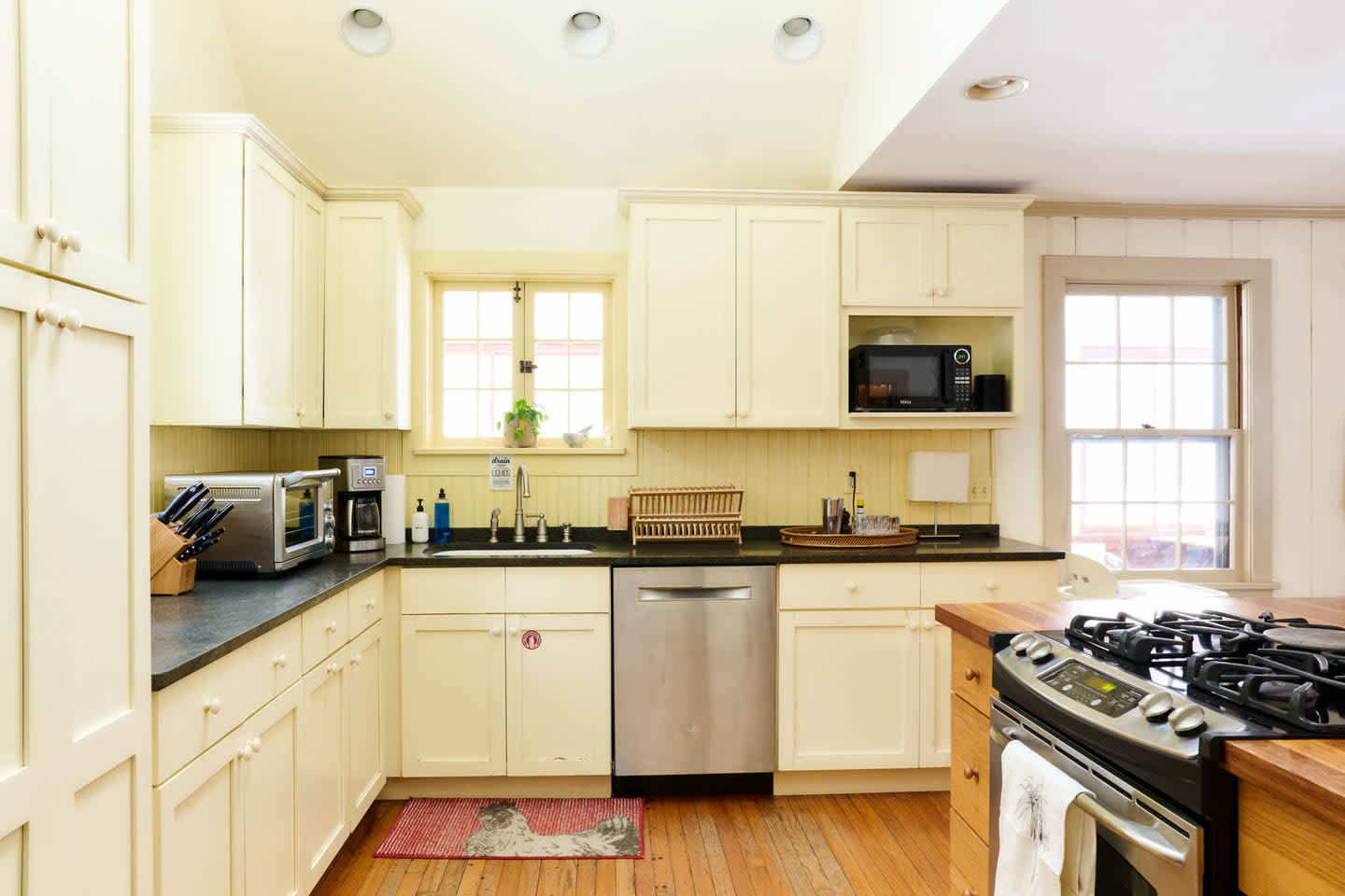 Bright kitchen with light yellow shaker-style cabinets, dark countertops, and stainless steel appliances including a dishwasher and microwave. A red rug adds a pop of color on the hardwood floor, and a window overlooks the sink area.