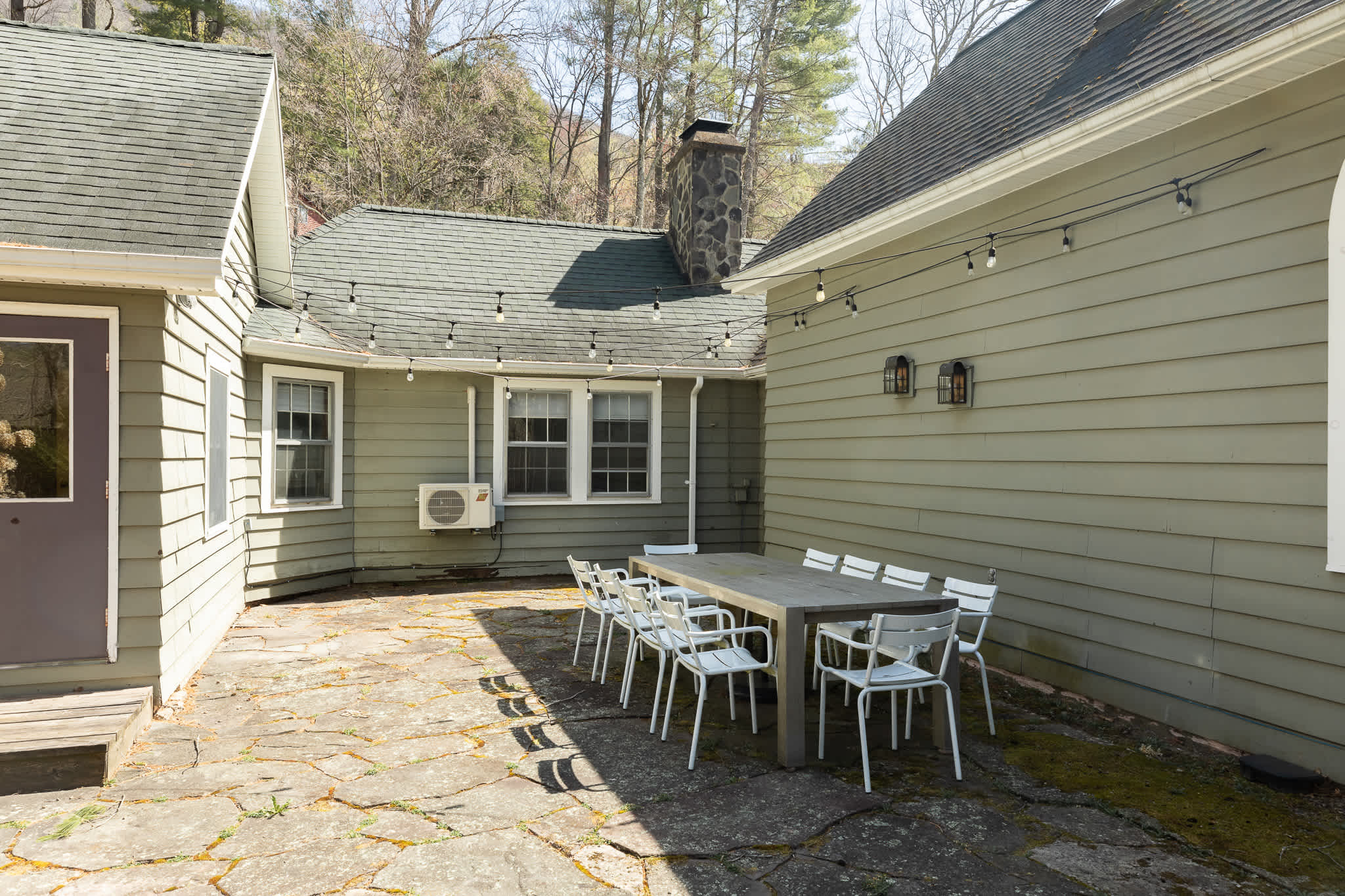 Outdoor flagstone patio adjacent to the house with green siding, featuring a long grey dining table and white chairs. String lights are strung overhead, and an outdoor AC unit is visible on the house wall.