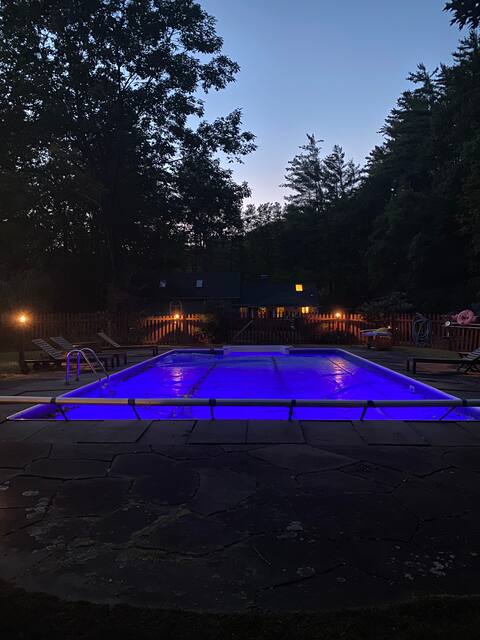 Nighttime view of a rectangular swimming pool, illuminated by vibrant blue underwater lights and covered by a safety cover. The pool is bordered by a stone patio, with dark silhouettes of surrounding trees and a distant house visible.